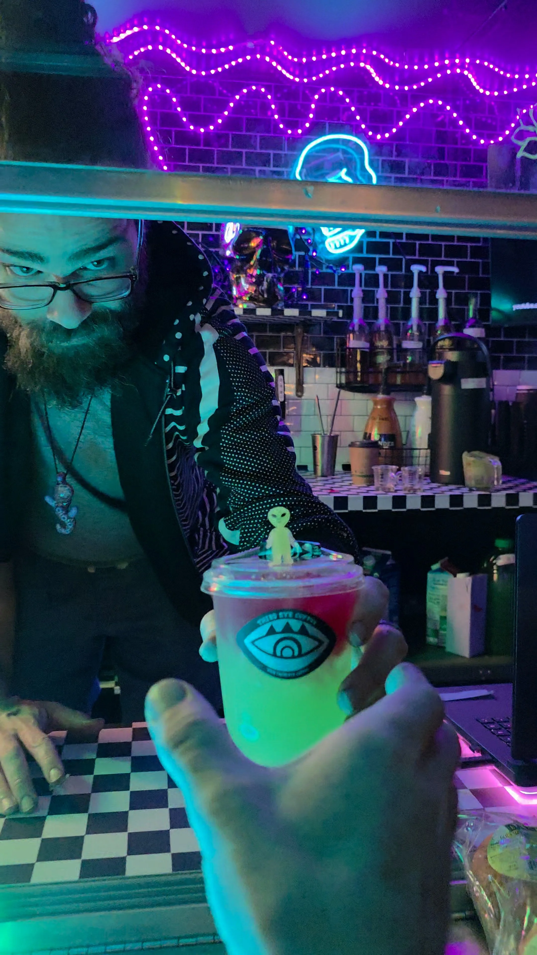 Person holding a drink with a small alien figure on top, in a neon-lit bar with psychedelic decor and a man behind the counter.