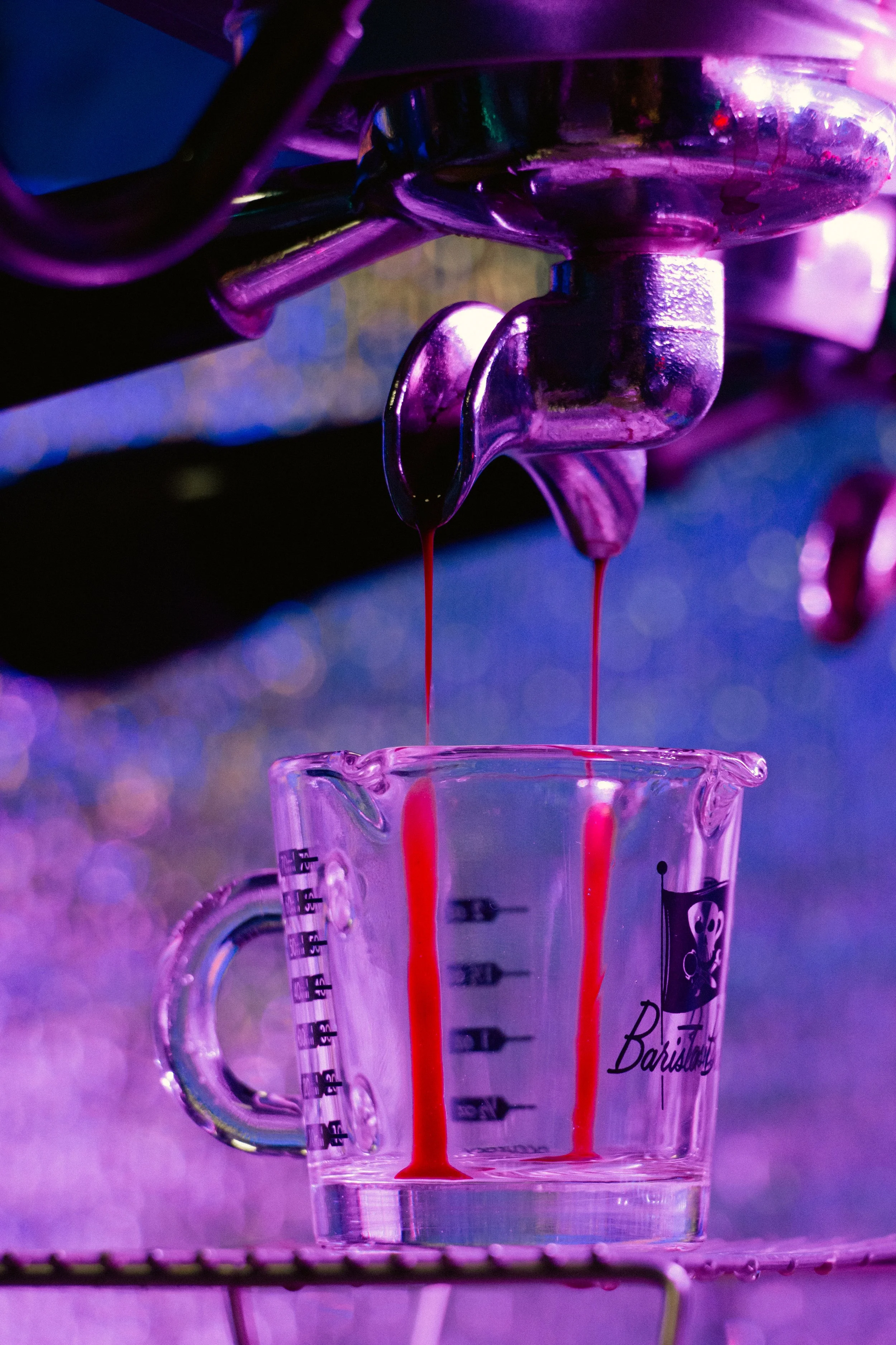 A close-up of a metallic valve with red liquid dripping into a clear measurement mug with purple and pink lighting.
