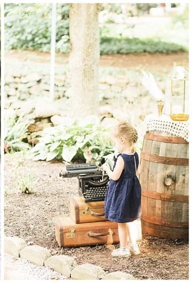 A young girl in a blue dress stands outdoors, next to a vintage typewriter placed on top of two stacked suitcases. There is a wooden barrel with a doily and decorative items nearby. The scene is set in a garden with trees and plants around.