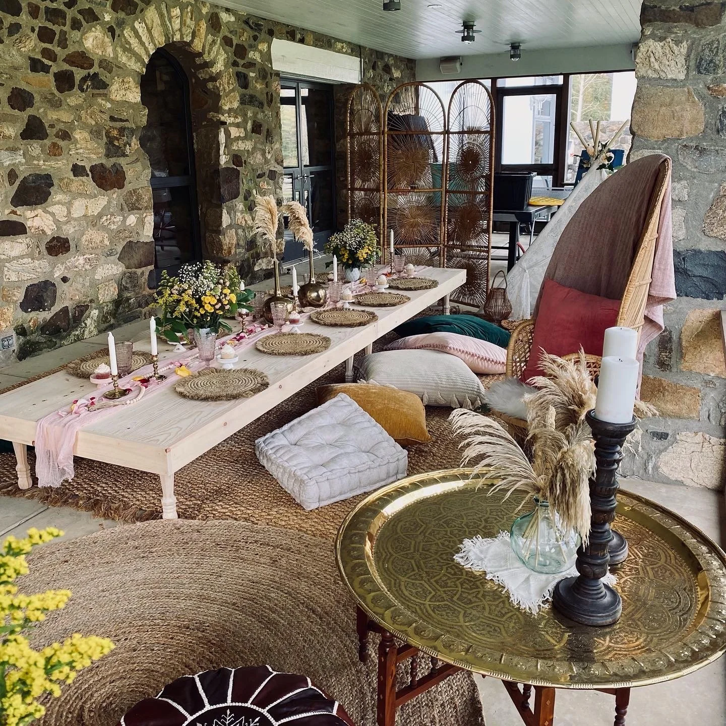 Rustic dining setup with wooden table and stone wall, featuring wicker chairs, candles, flowers, and cushions.