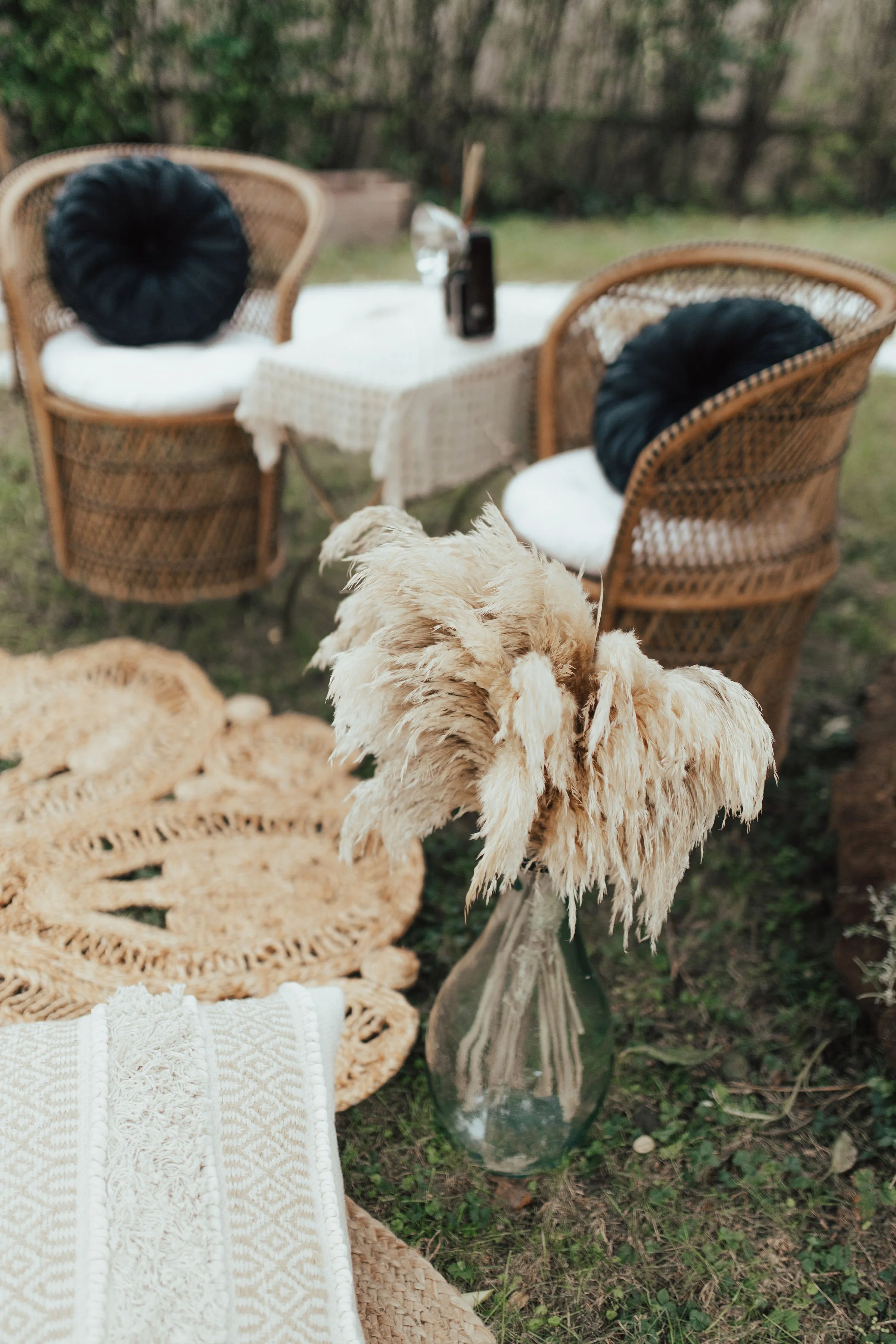 Outdoor bohemian seating area with wicker chairs, round crocheted rug, pampas grass in a glass vase, and patterned pillow on grass.