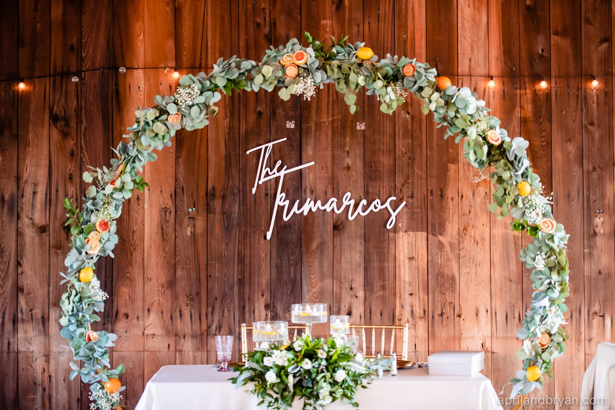 Wedding backdrop with wooden wall, circular greenery arch with citrus accents, and "The Trimarcos" sign in white script. Table with floral centerpiece and candles.