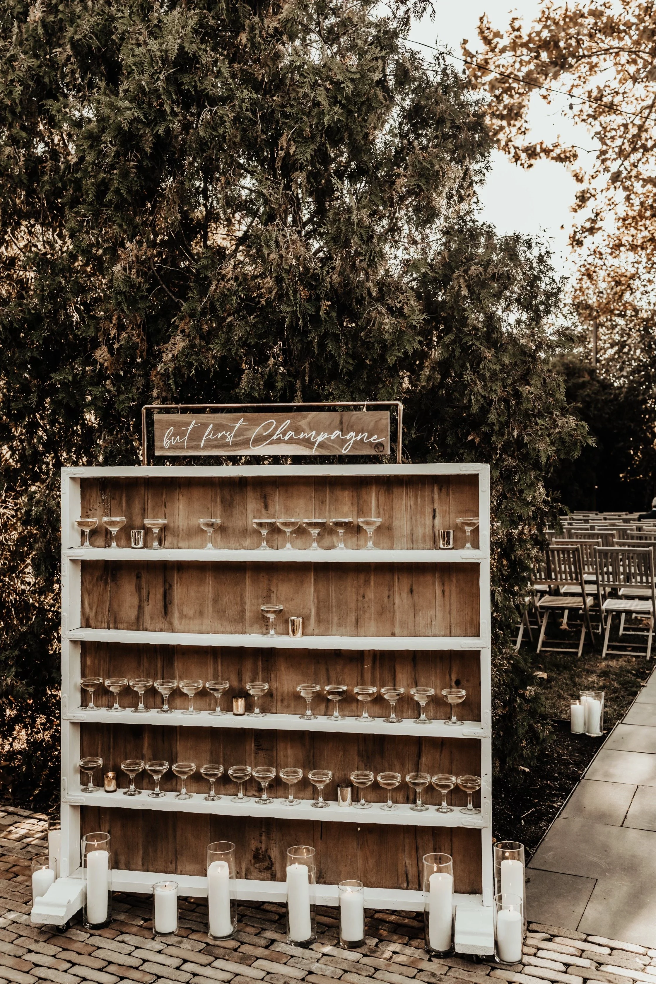 Wooden display with champagne glasses and candles, sign reads 'but first, champagne,' outdoor setting.