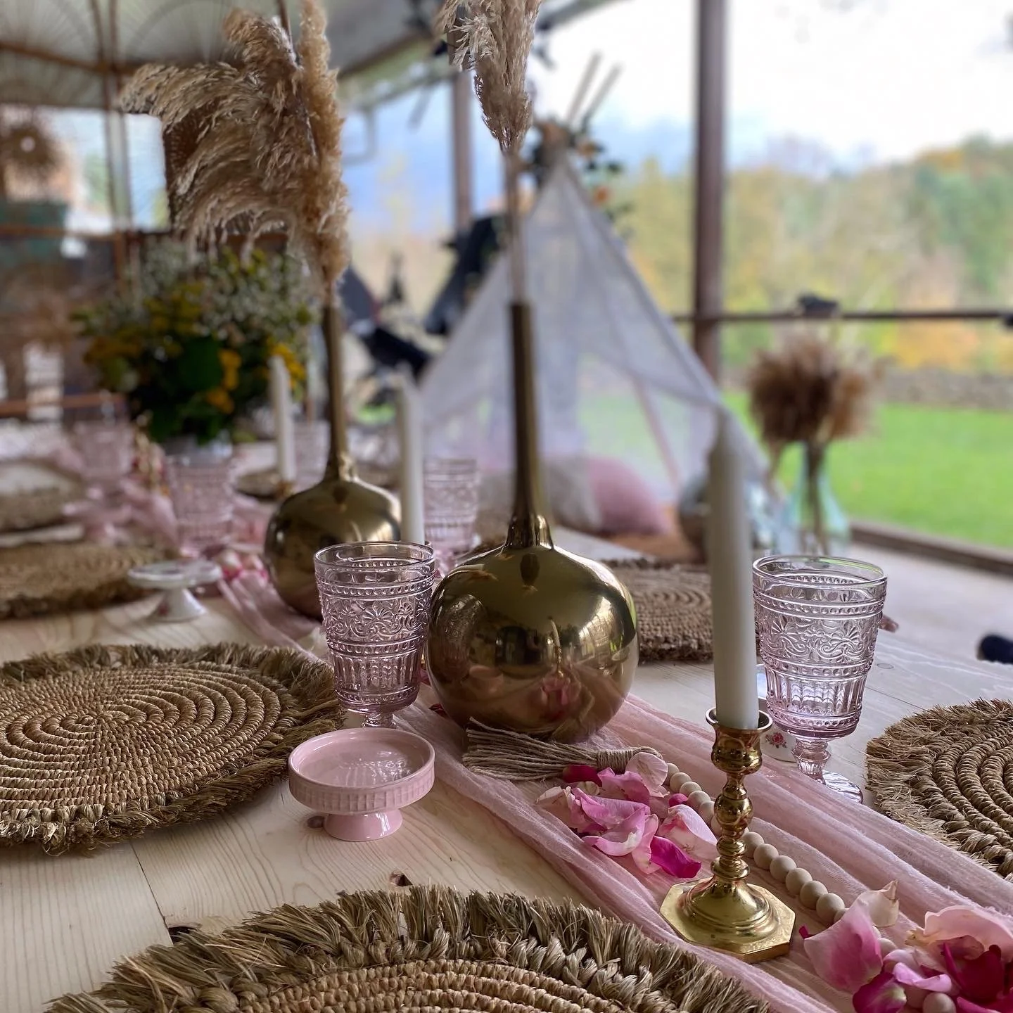 Elegant table setting with decorative woven placemats, golden candle holders and vases, glassware, and floral accents on light wooden table.
