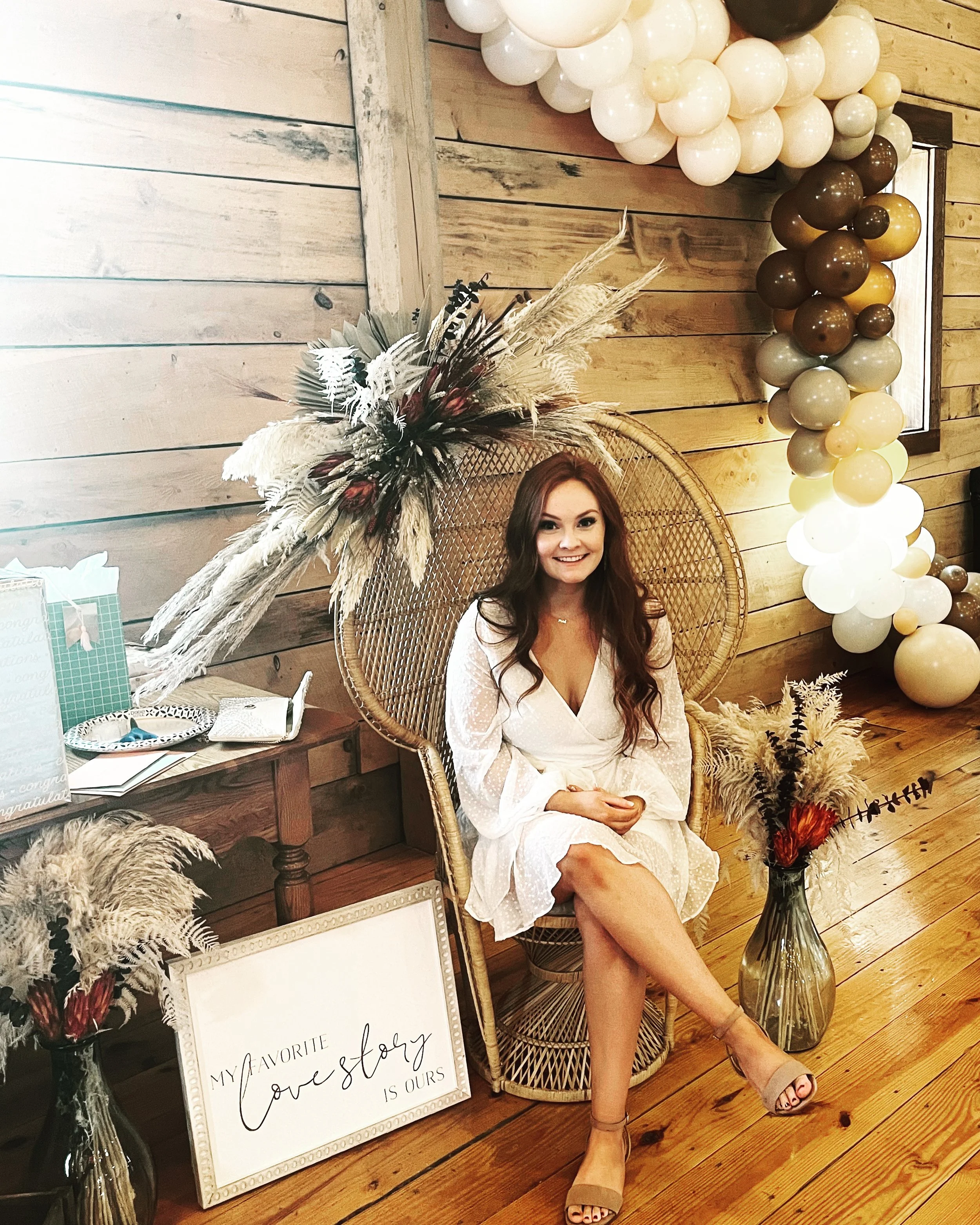 Woman sitting in a wicker chair, surrounded by rustic decor including dried flowers in vases, a sign reading "My favorite love story is ours," and a balloon arch. She wears a white dress and is in a wood-paneled room.