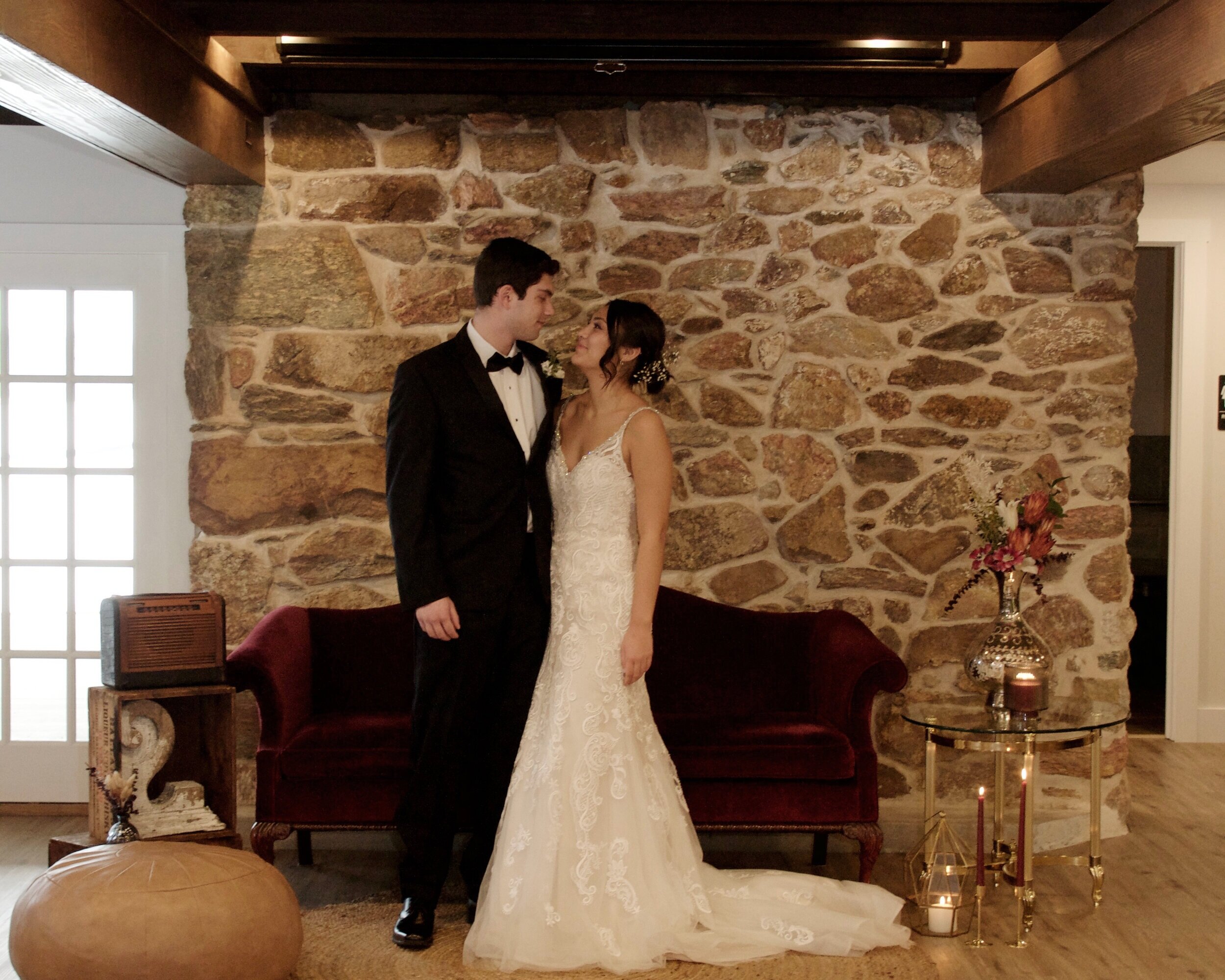 A bride and groom standing in formal attire, smiling at each other, in front of a rustic stone wall. The bride is in a white lace gown, and the groom is in a black tuxedo. They are standing near a red velvet couch with decorative items around, includ
