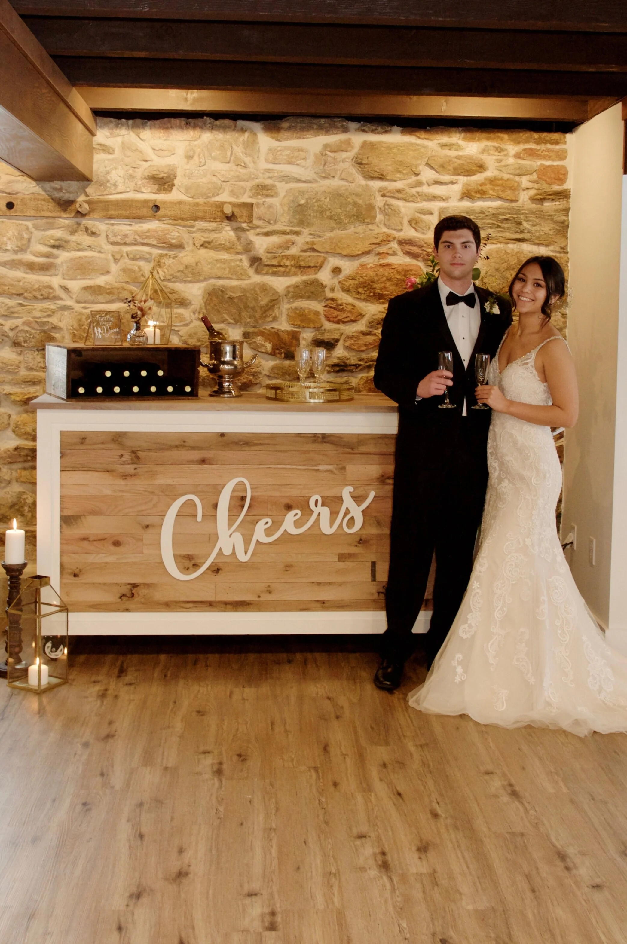 A bride and groom standing by a bar with a sign that says "Cheers." The bride is in a lace wedding dress, and the groom is in a tuxedo. The bar has decorative items and a stone wall backdrop.