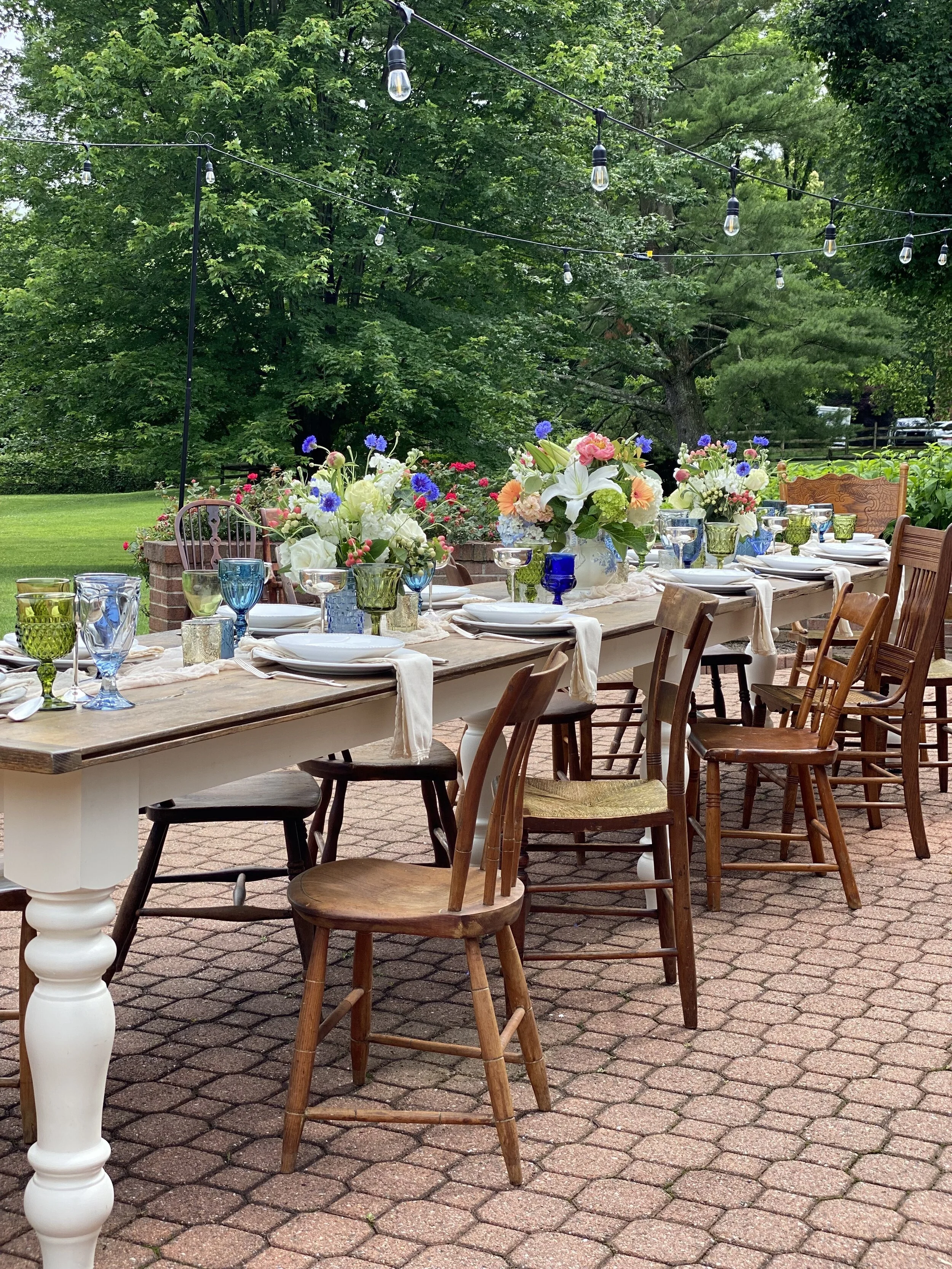 Outdoor table setting on a brick patio, featuring a long wooden table with mismatched chairs, set with colorful glassware, white plates, and floral centerpieces. String lights hang above, and lush greenery surrounds the area.