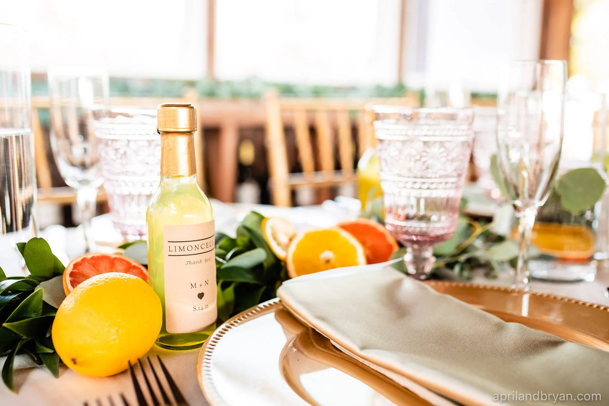 Table setting with a bottle of Limoncello, sliced citrus fruits, glassware, and a gold-rimmed plate with a folded napkin.