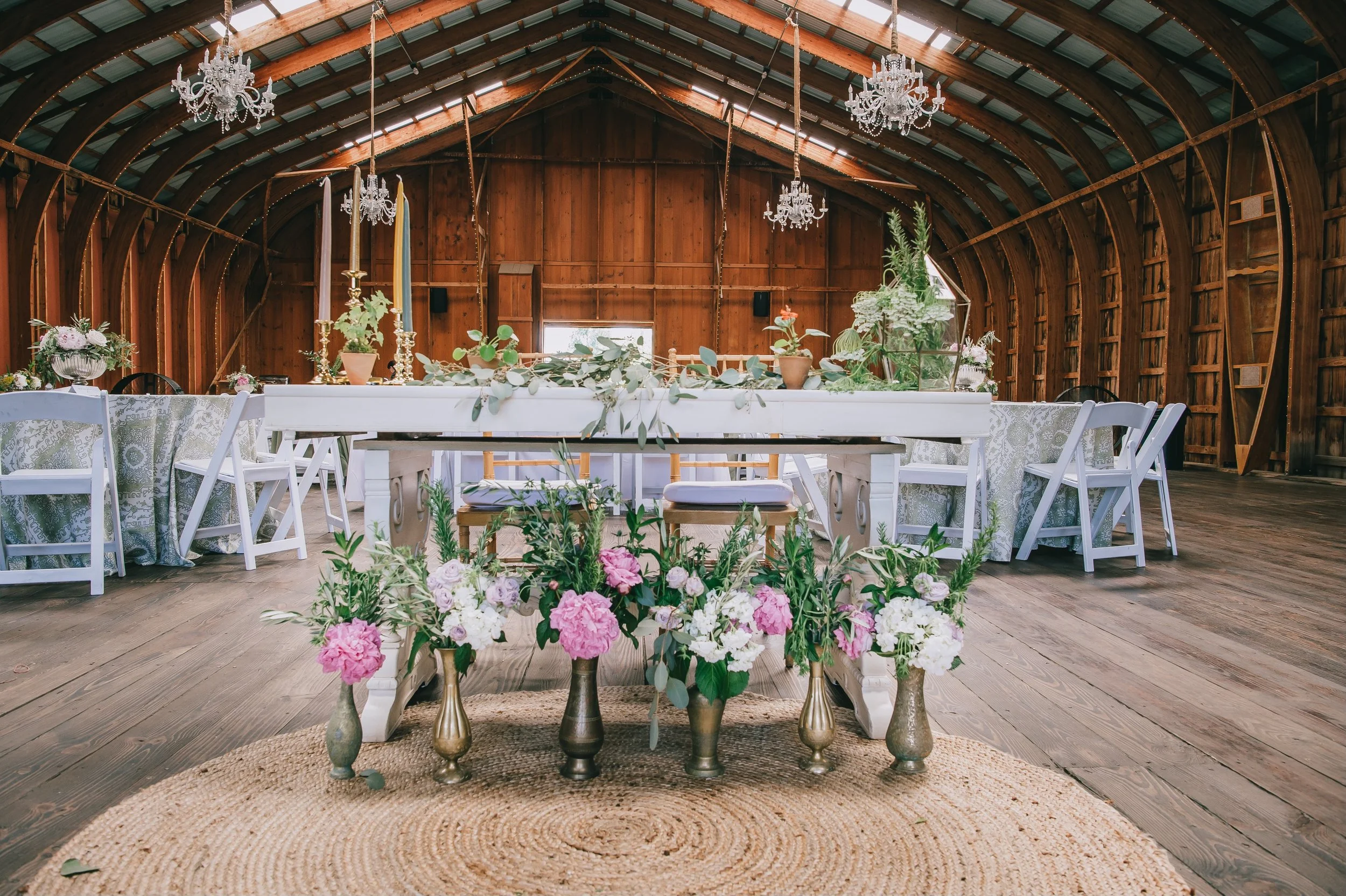 A rustic barn-style venue decorated for an event with wooden interior and chandeliers. A white table is adorned with plants and gold candelabras, surrounded by white chairs. Several flower vases with pink and white blooms are arranged on a round wove