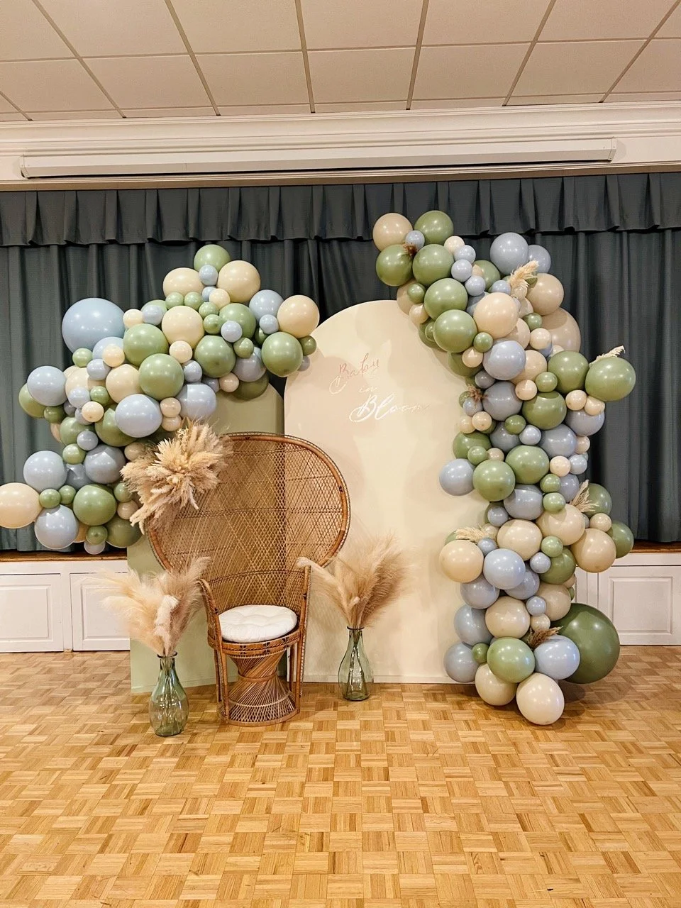 Decorative backdrop with green, beige, and light blue balloons, a wicker chair with a cushion, pampas grass in glass vases, and a wooden floor.