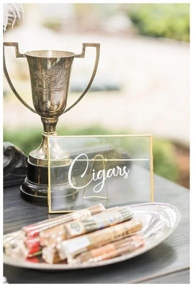 A table with a trophy, a glass sign labeled "Cigars," and a silver tray holding assorted cigars.