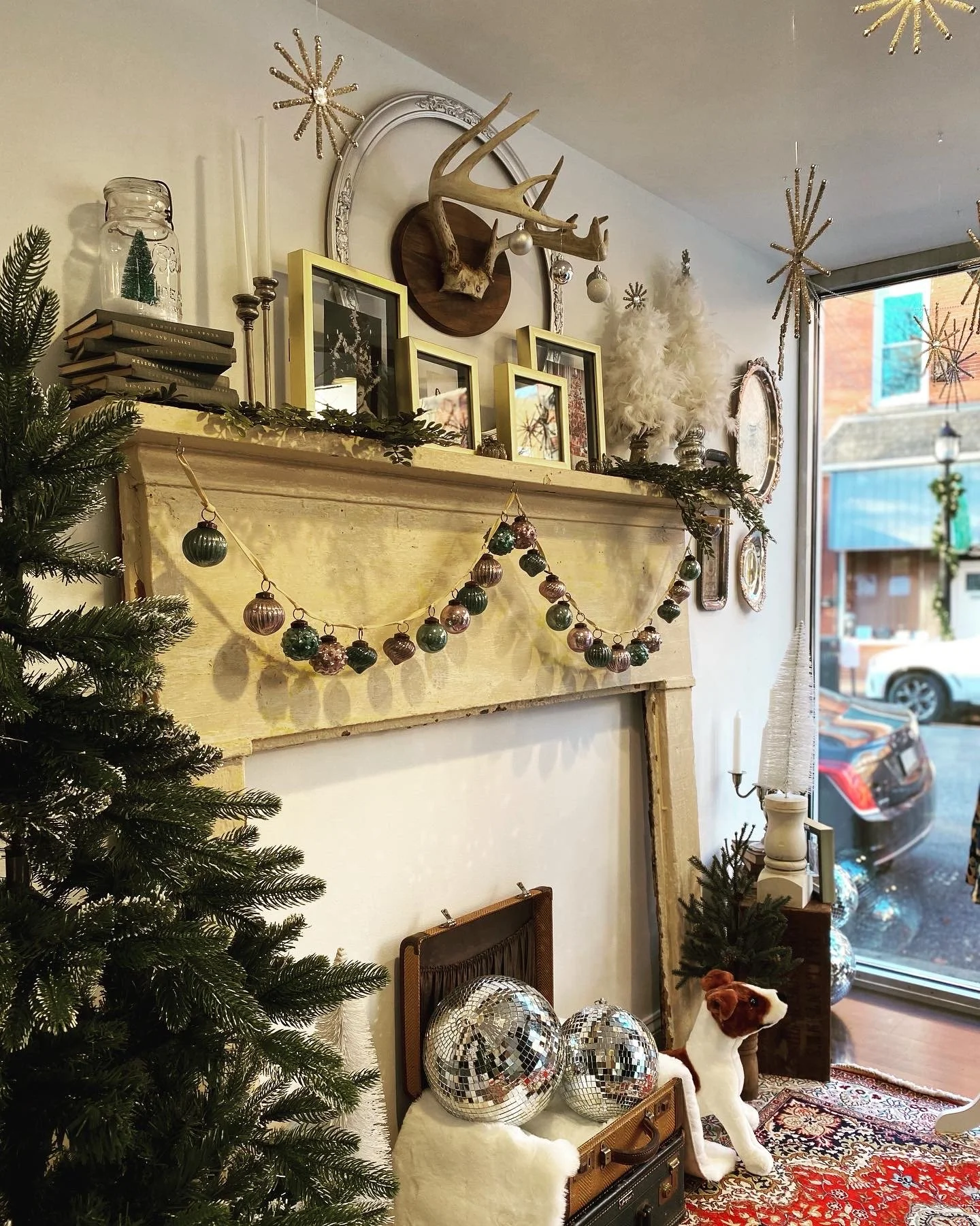 Holiday-themed interior decoration featuring a mantelpiece adorned with garlands, ornaments, antlers, framed photos, tall candles, and a vintage jar. Below the mantel are disco balls in a suitcase, a plush dog toy, and a small Christmas tree. Additio