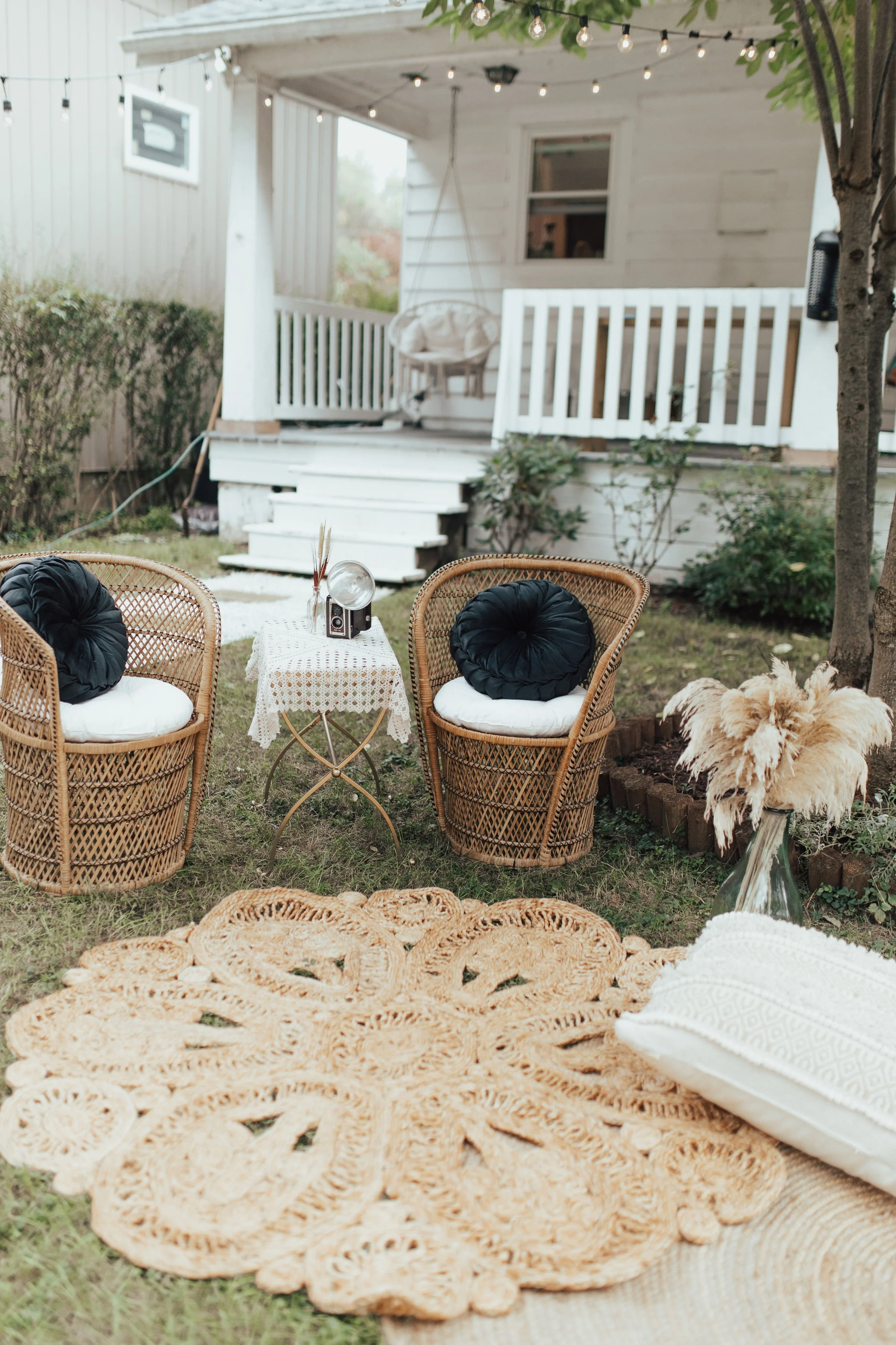 Bohemian-style outdoor seating area with two wicker chairs, black cushions, a small round table with a lace tablecloth, an old-fashioned camera, and decorative pampas grass. There are string lights above, a woven rug on the grass, and a white porch i