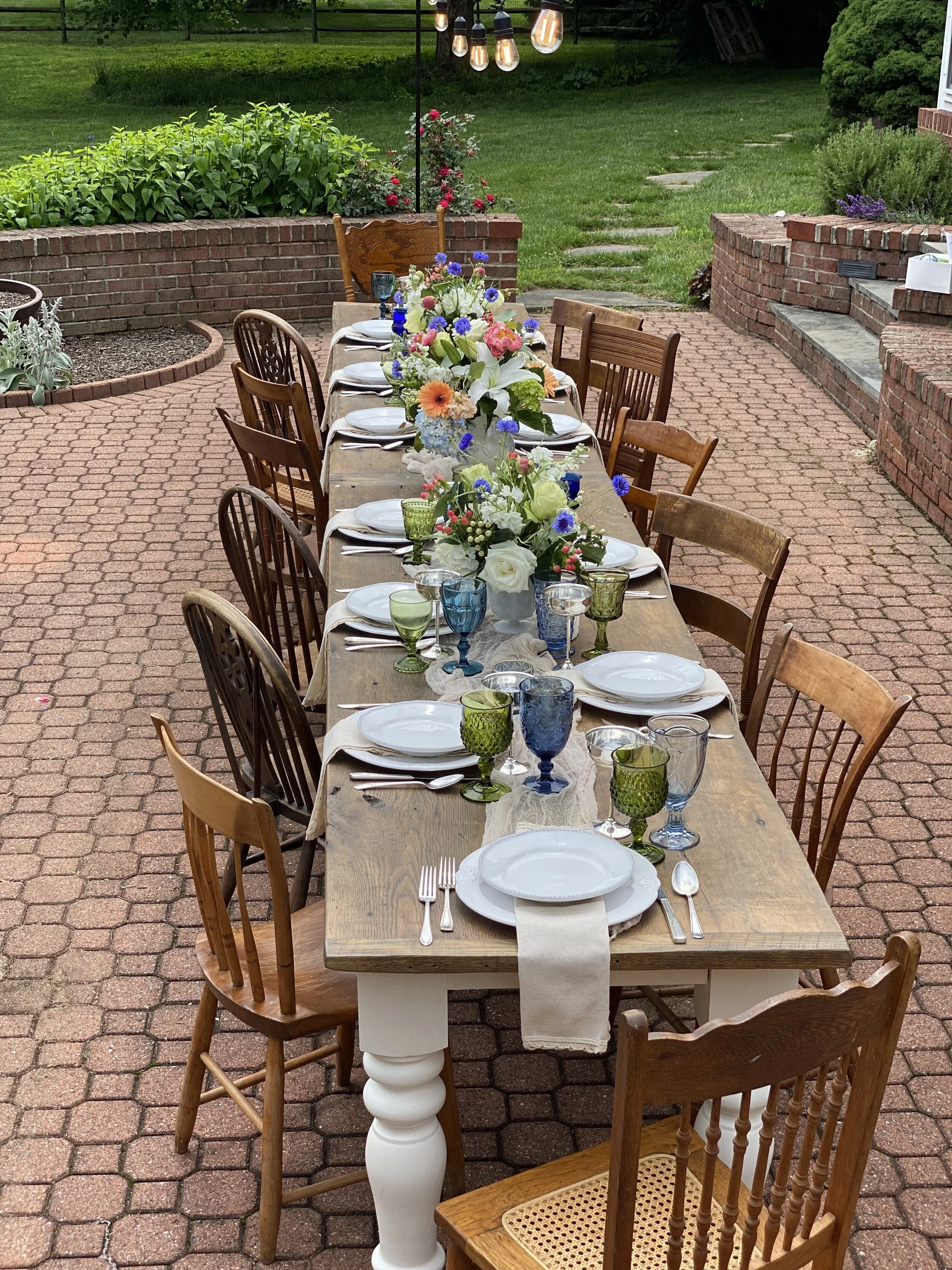 Outdoor dining table set for a meal with mixed chairs, white tablecloth, floral centerpiece, and colorful glassware.