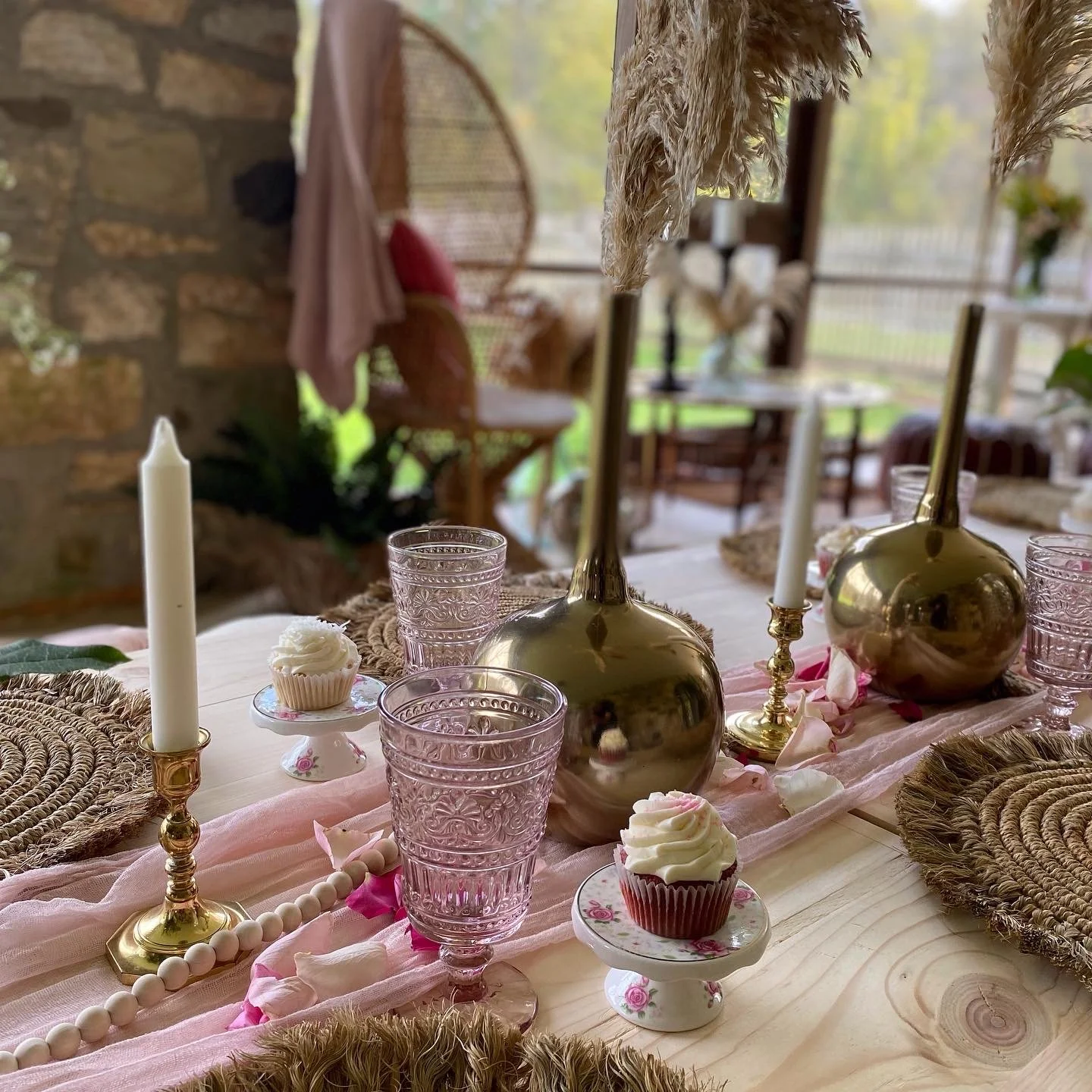Rustic table setting with pink glasses, cupcakes on floral plates, gold candlesticks, pampas grass in vases, and woven placemats on a wooden surface.