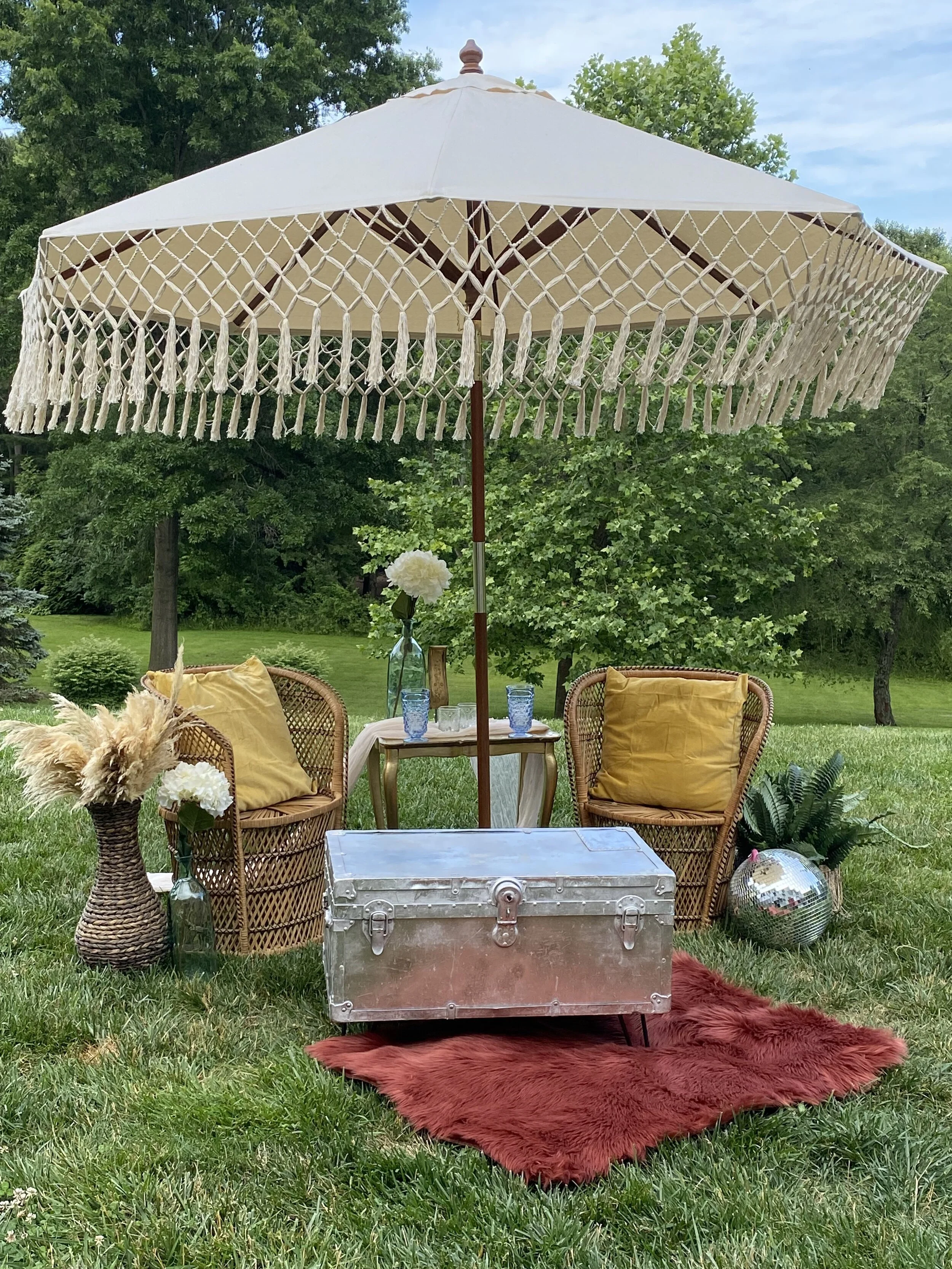 Outdoor seating arrangement with wicker chairs, fringe parasol, metallic chest, and decorative items on grass.