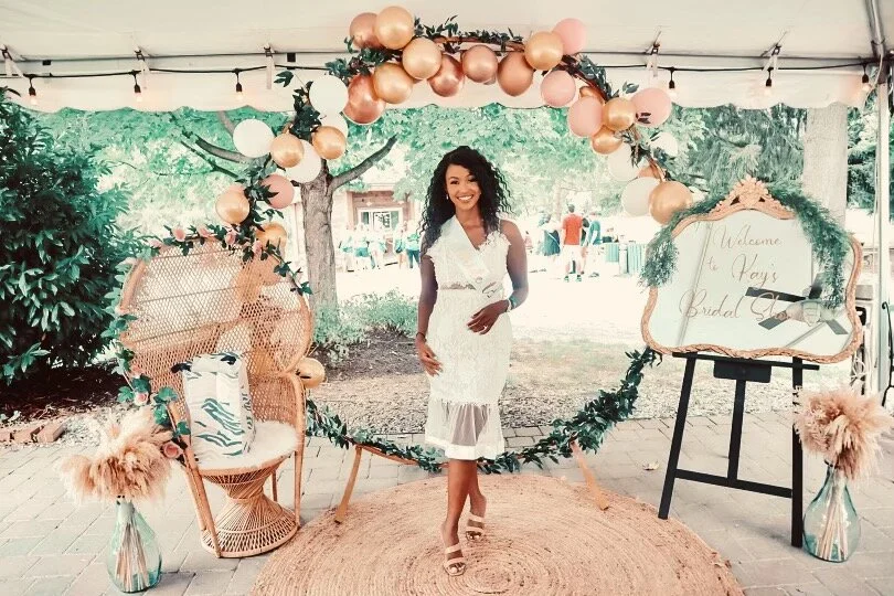 Woman standing at a bridal shower, surrounded by a decorative arch with balloons and greenery, next to a wicker chair and a welcome sign.