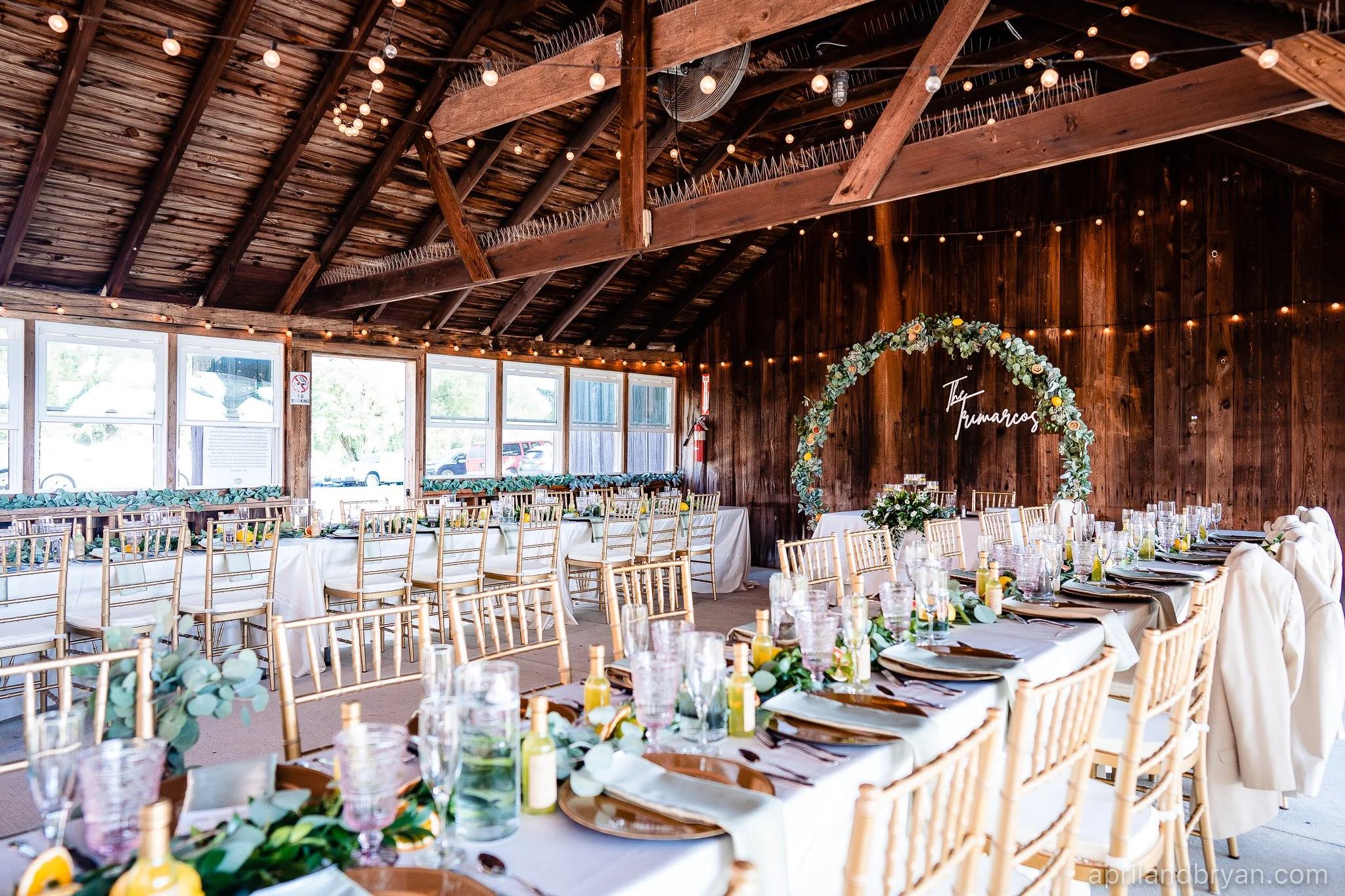 Rustic wedding reception with wooden beams, string lights, long tables with white linens, gold chairs, eucalyptus decorations, and a floral arch.