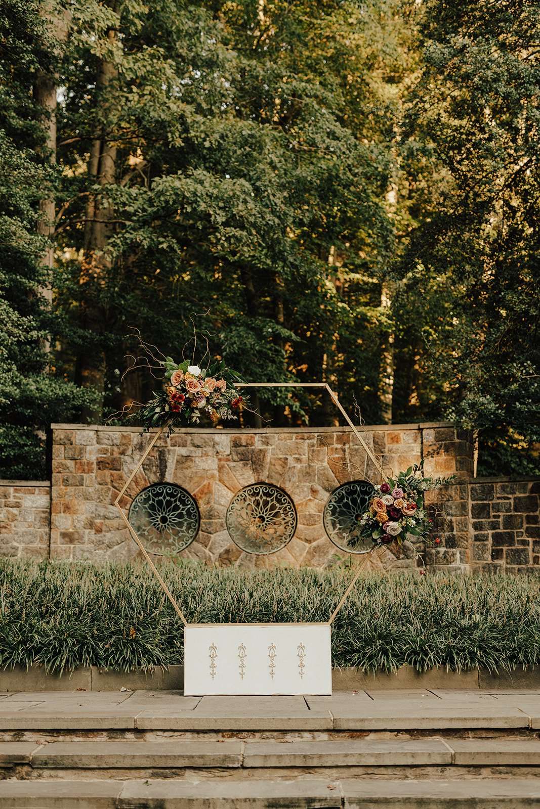 Wedding ceremony setup with geometric arch, floral arrangements, and stone wall in outdoor garden setting.