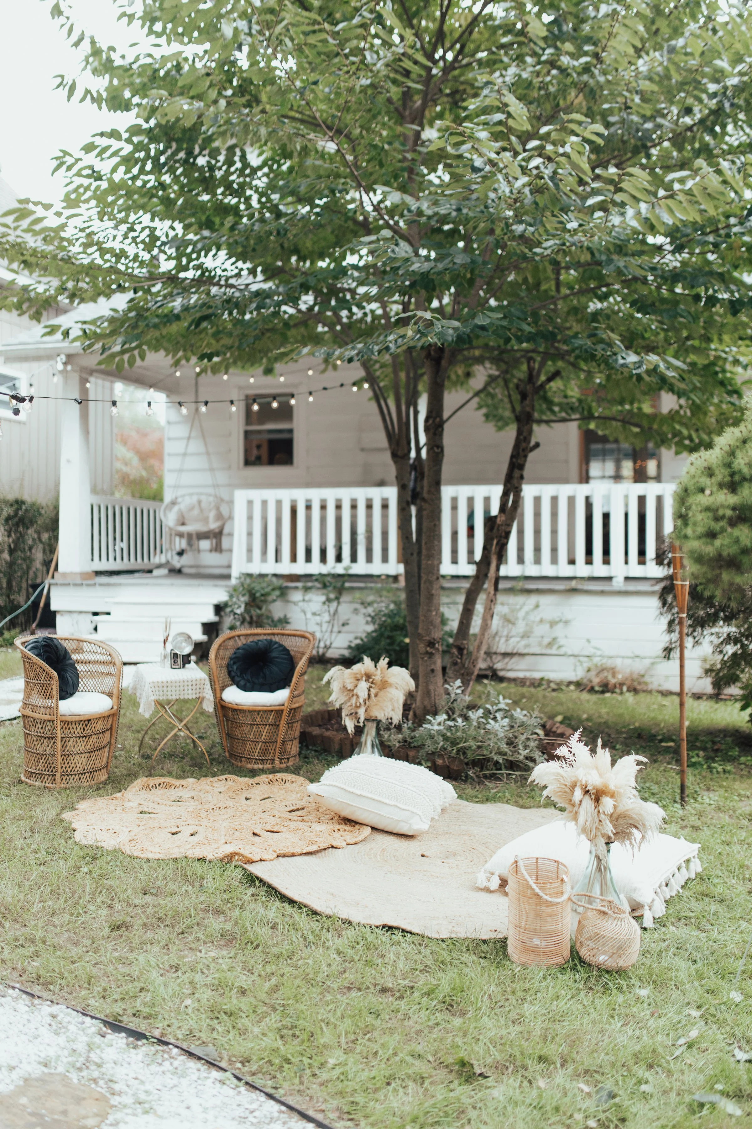 Outdoor seating area with wicker chairs and cushions on rugs, under a tree, near a porch. Decor includes string lights and dried flowers in vases.