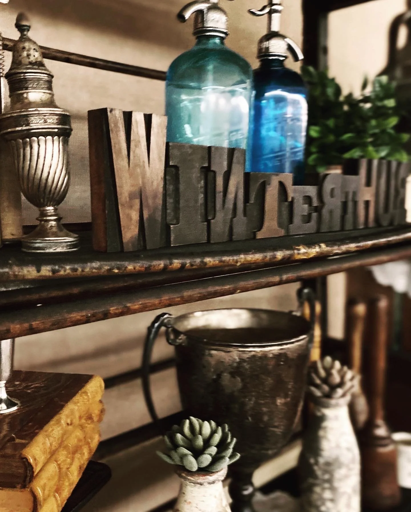Vintage display with decorative silver urn, glass bottles, wooden letters spelling 'WINTERTHUR', potted succulents, stacked books, and a metallic bucket on a shelf.