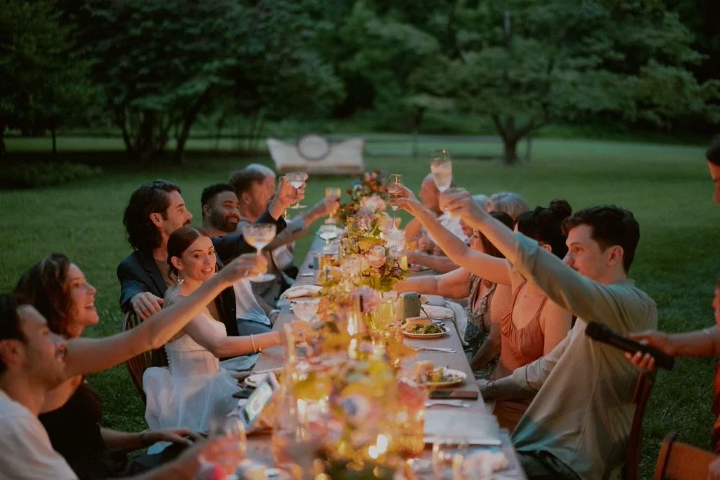 People sitting at a long outdoor table toasting with drinks, surrounded by greenery, bouquets, and evening lighting.