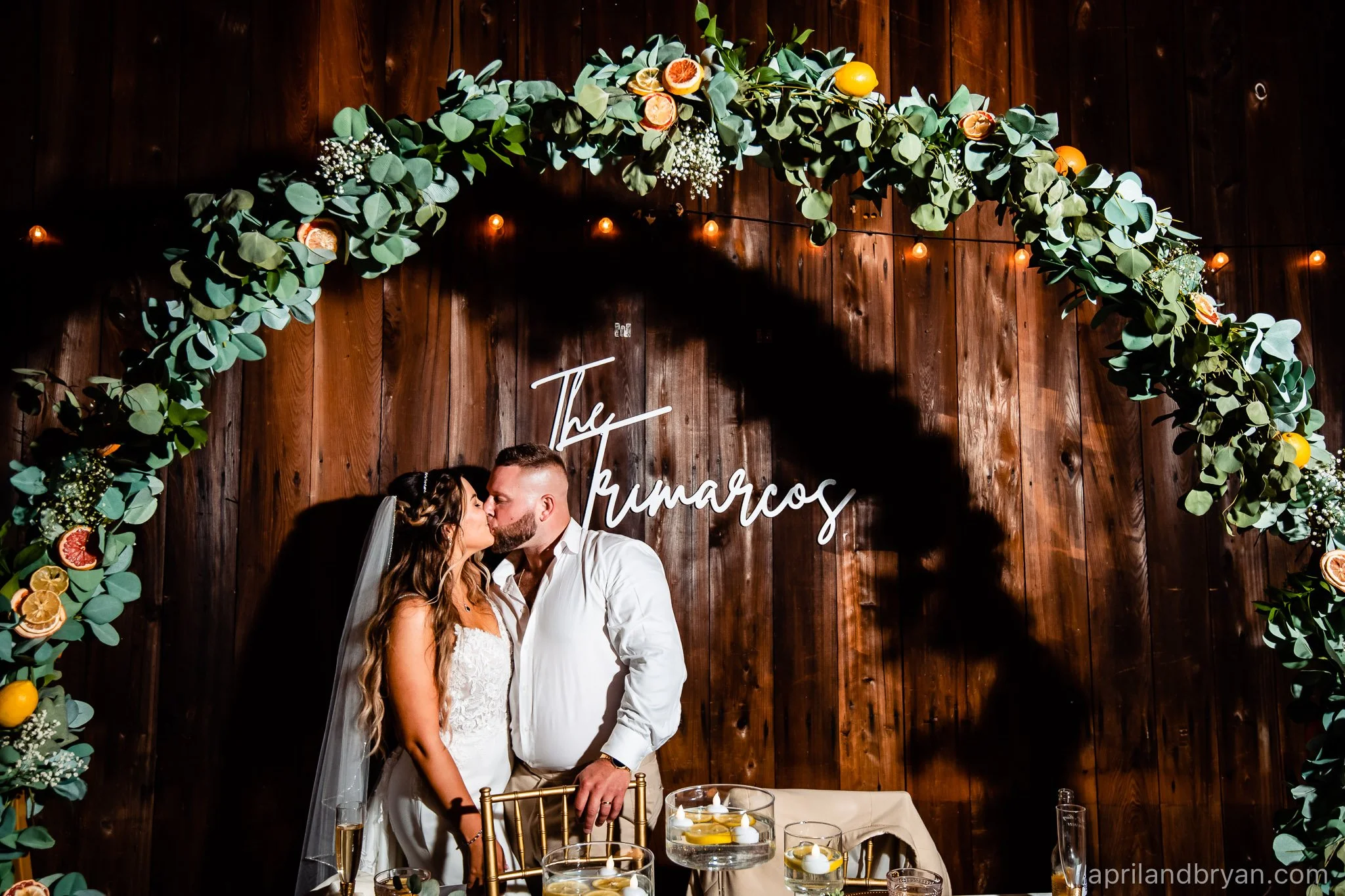 Wedding couple kissing under a floral arch with a wooden backdrop and illuminated sign.