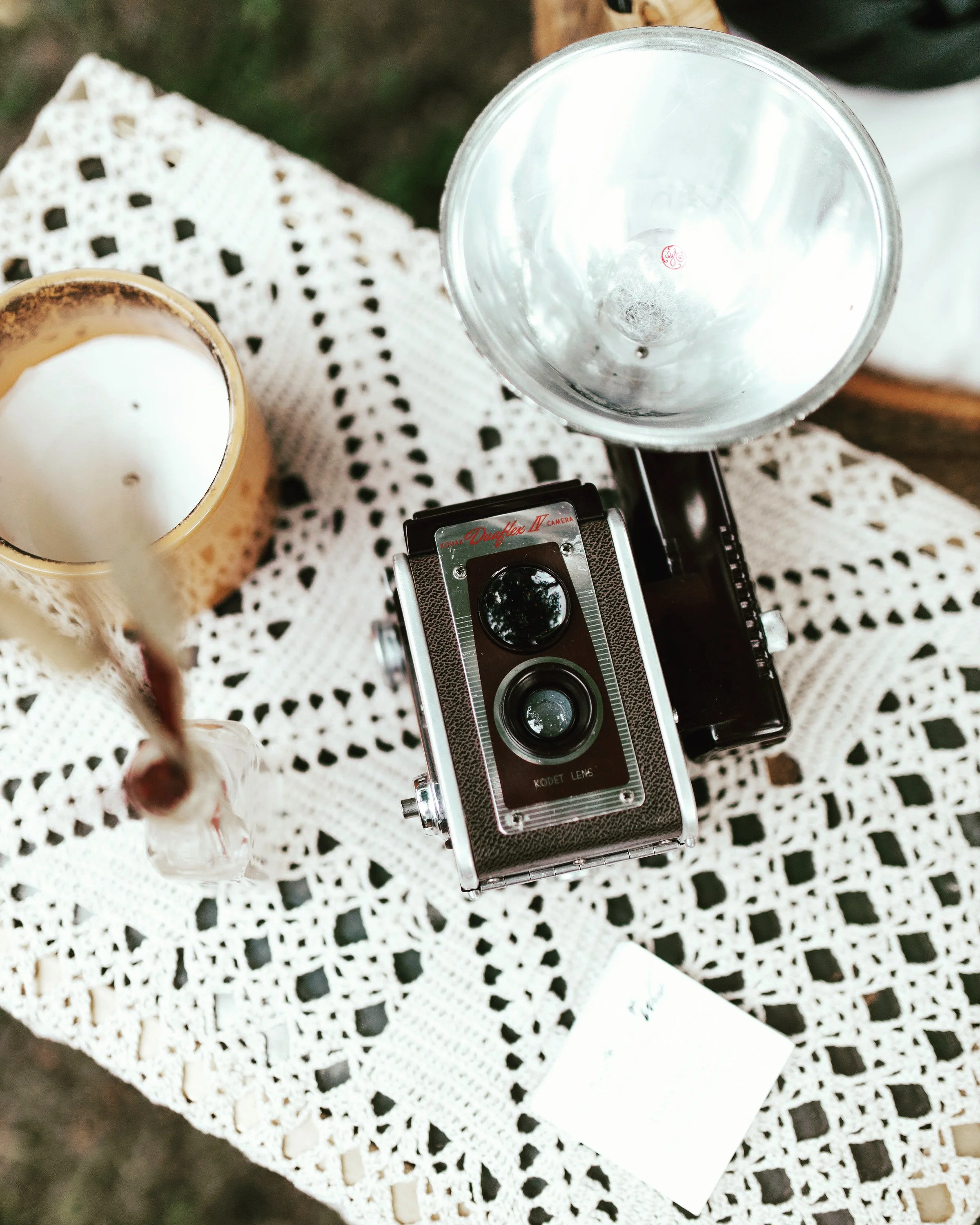 Vintage camera on a crochet tablecloth with a candle, vase, and business card nearby.