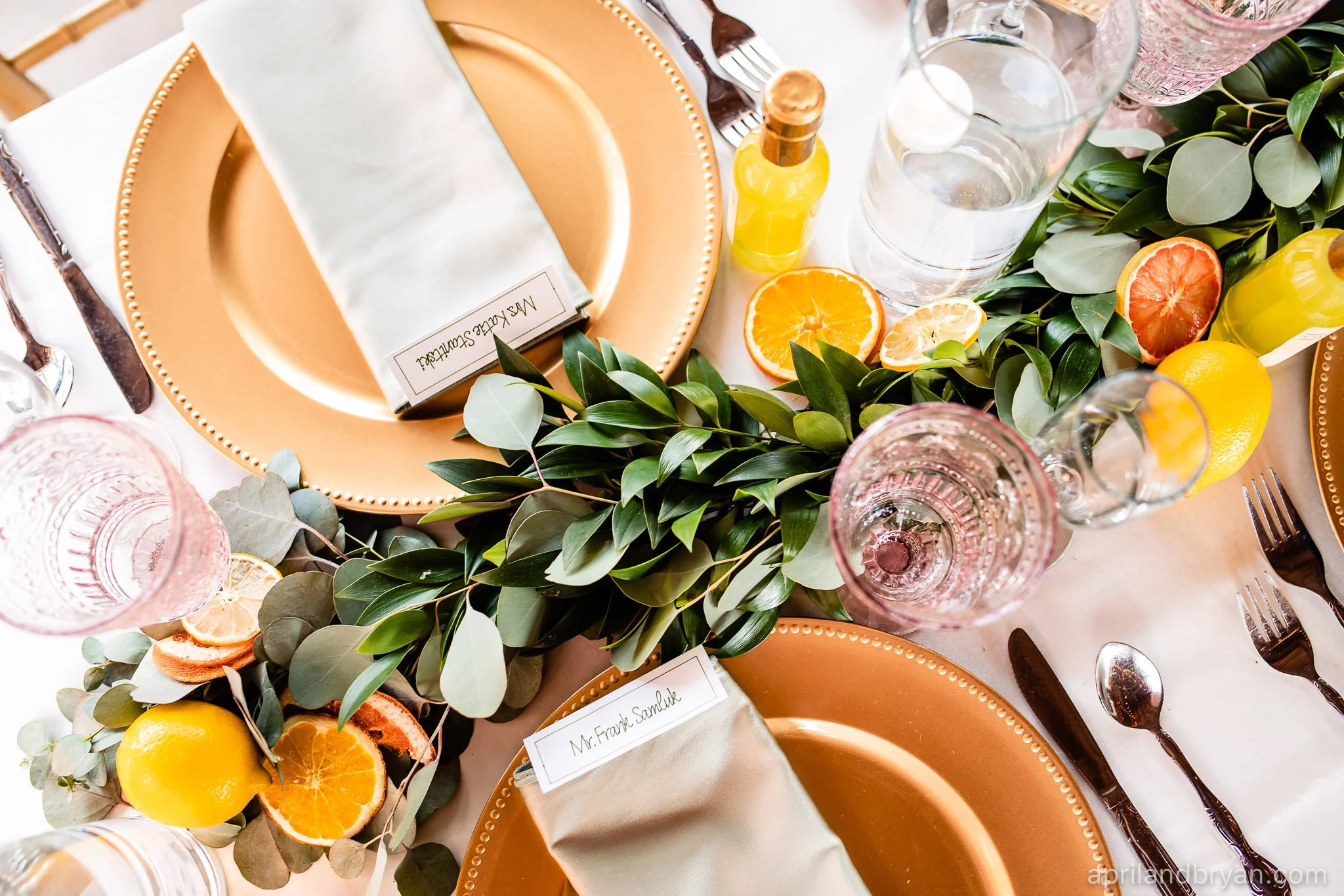 Elegant table setting with gold plates, green foliage, sliced citrus fruits, glassware, and cutlery.