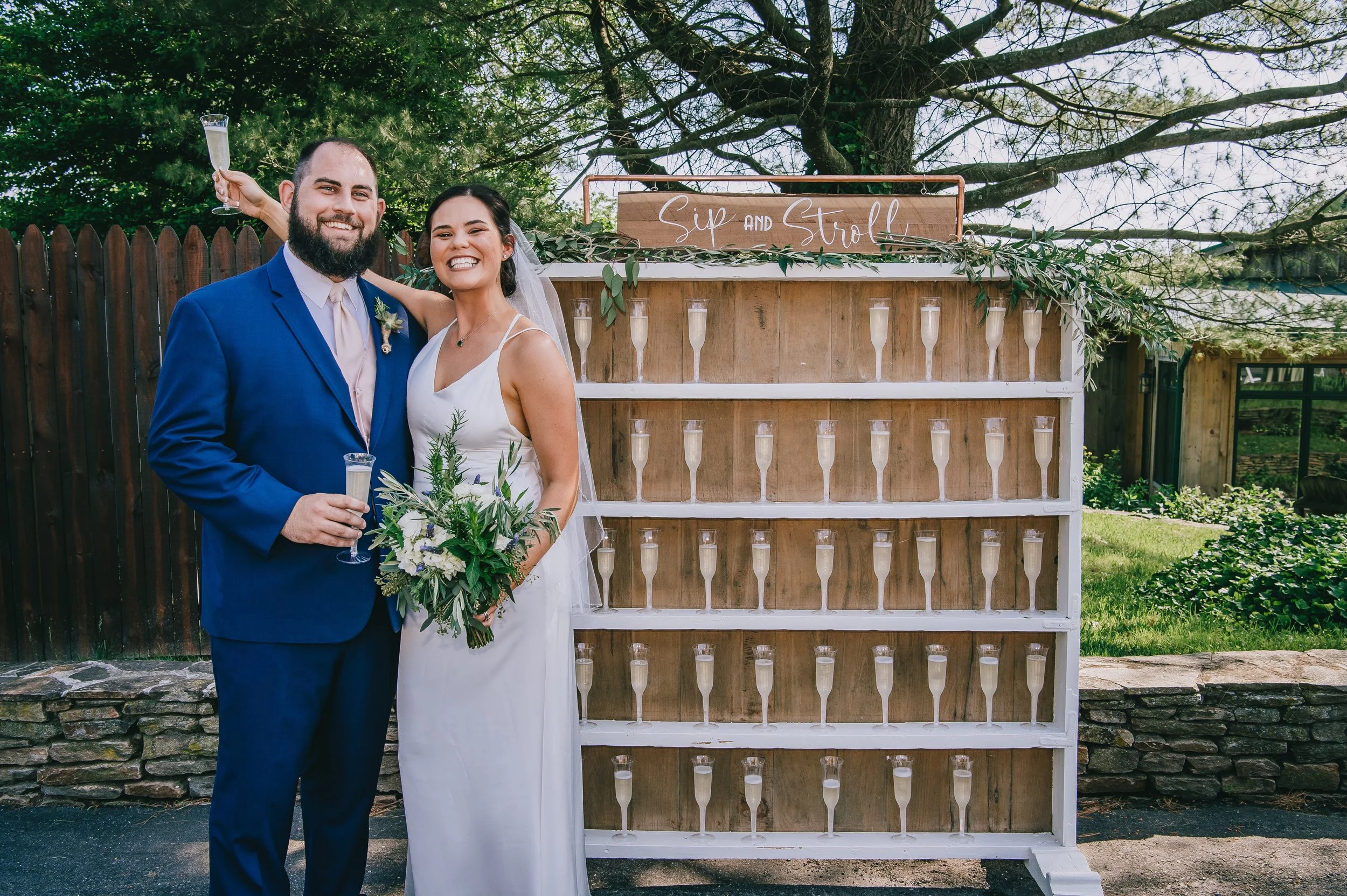 Wedding couple smiling in front of a champagne shelf labeled 'Sip and Stroll,' holding glasses of champagne, with greenery and outdoor setting in the background.