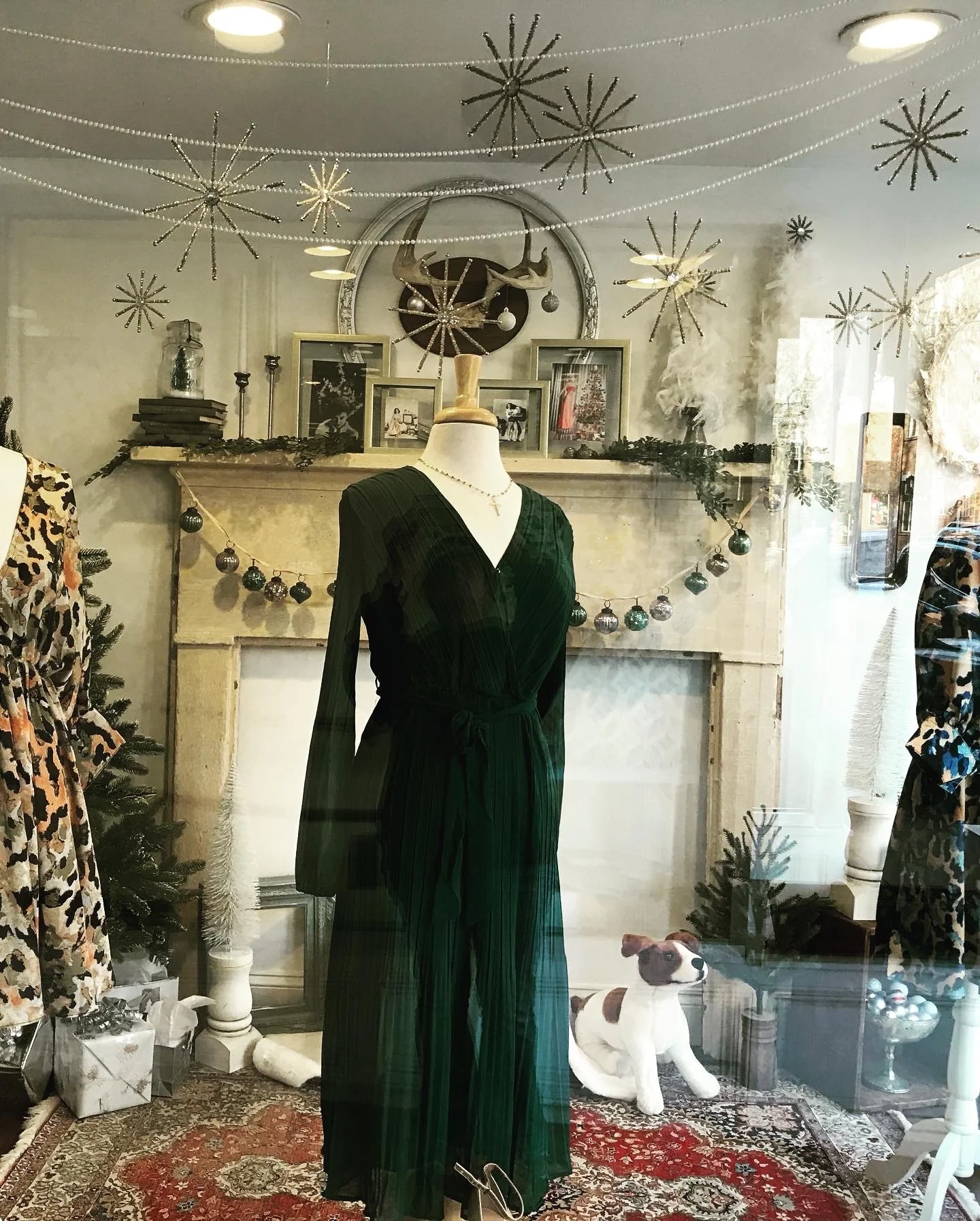 Store window display featuring a mannequin with a green dress, surrounded by festive decorations like snowflakes and ornaments, a faux dog, and framed photos on a mantelpiece.
