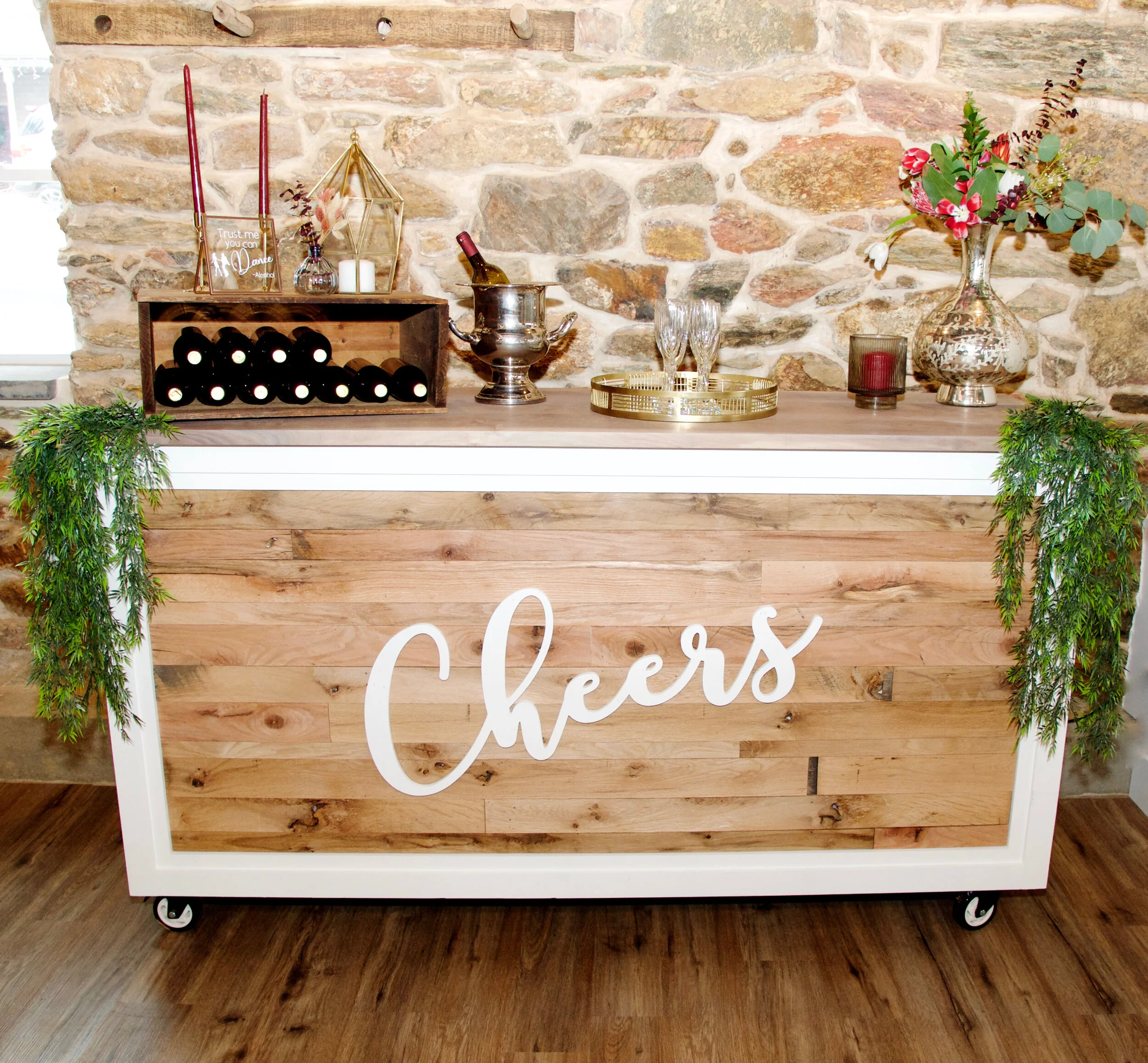 Rustic bar cart with "Cheers" sign, wine bottles, glasses, candle, and floral decor against a stone wall.