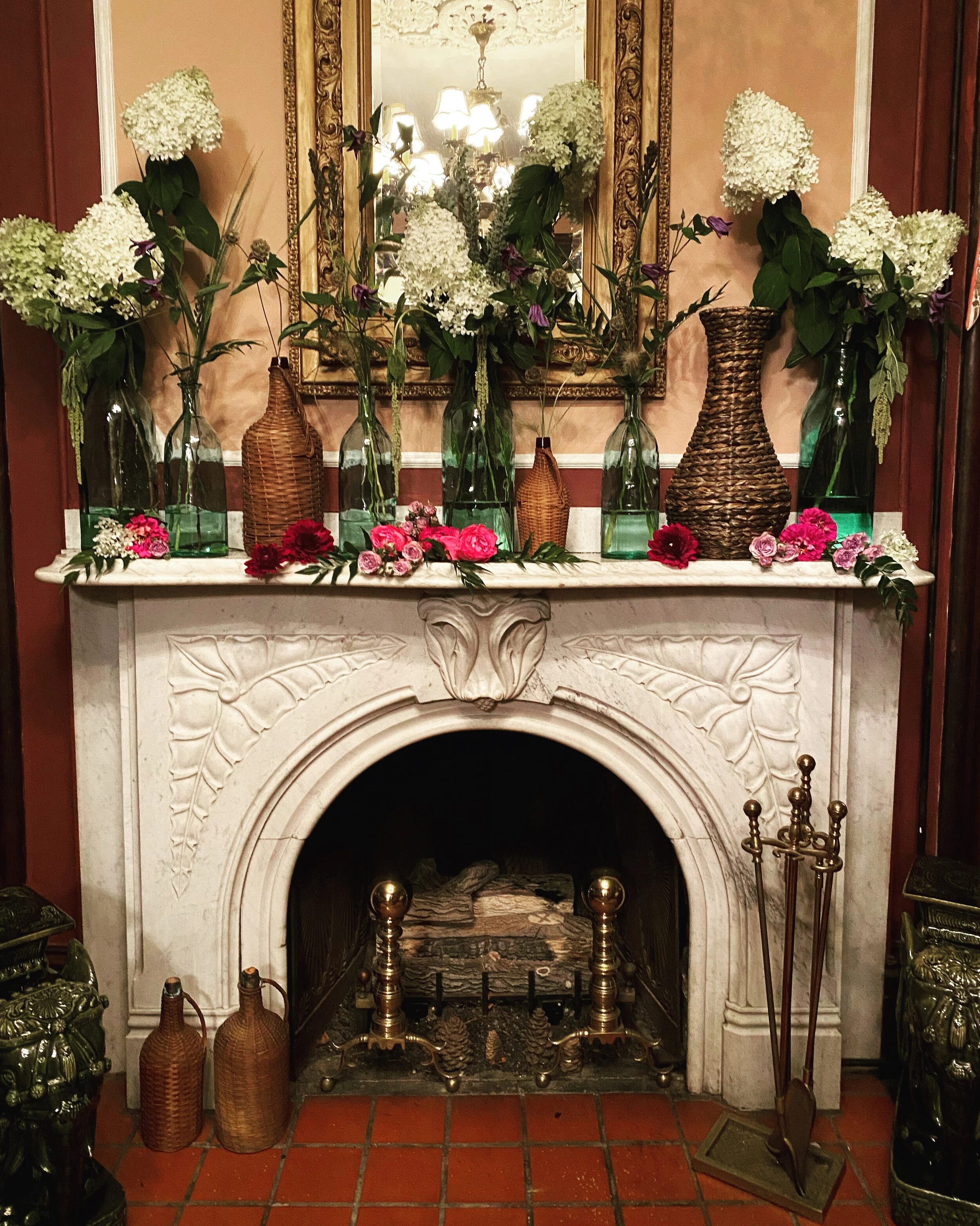 A fireplace with a white marble mantel, decorated with tall glass vases and wicker baskets filled with white and pink flowers and greenery, a large ornate mirror above, and brass fireplace tools on the side.