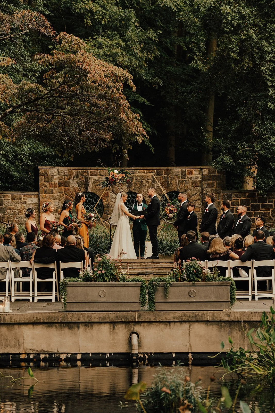Outdoor wedding ceremony surrounded by lush greenery; bride and groom standing at the altar with officiant; bridesmaids and groomsmen beside them; guests seated in white chairs.