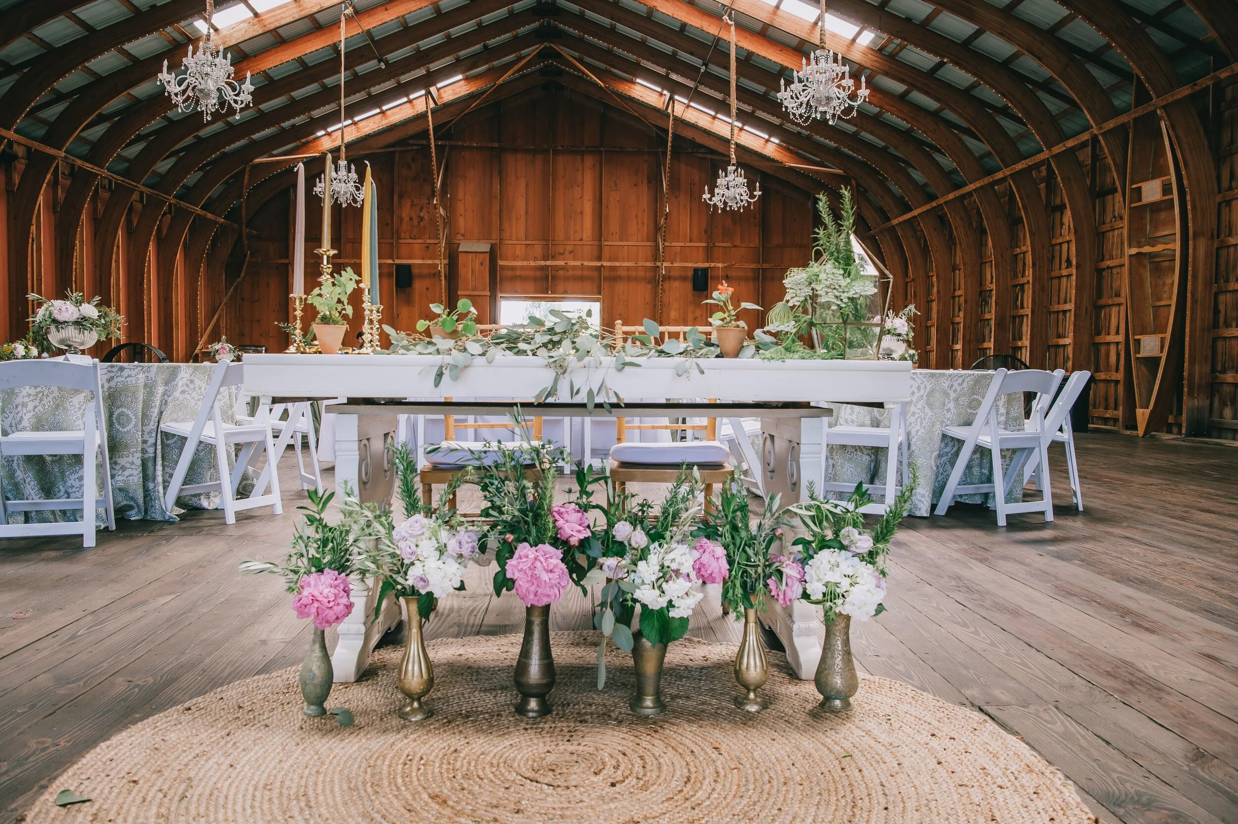 Rustic barn interior decorated for an event with chandeliers, white wooden tables, floral arrangements in brass vases, and white folding chairs.