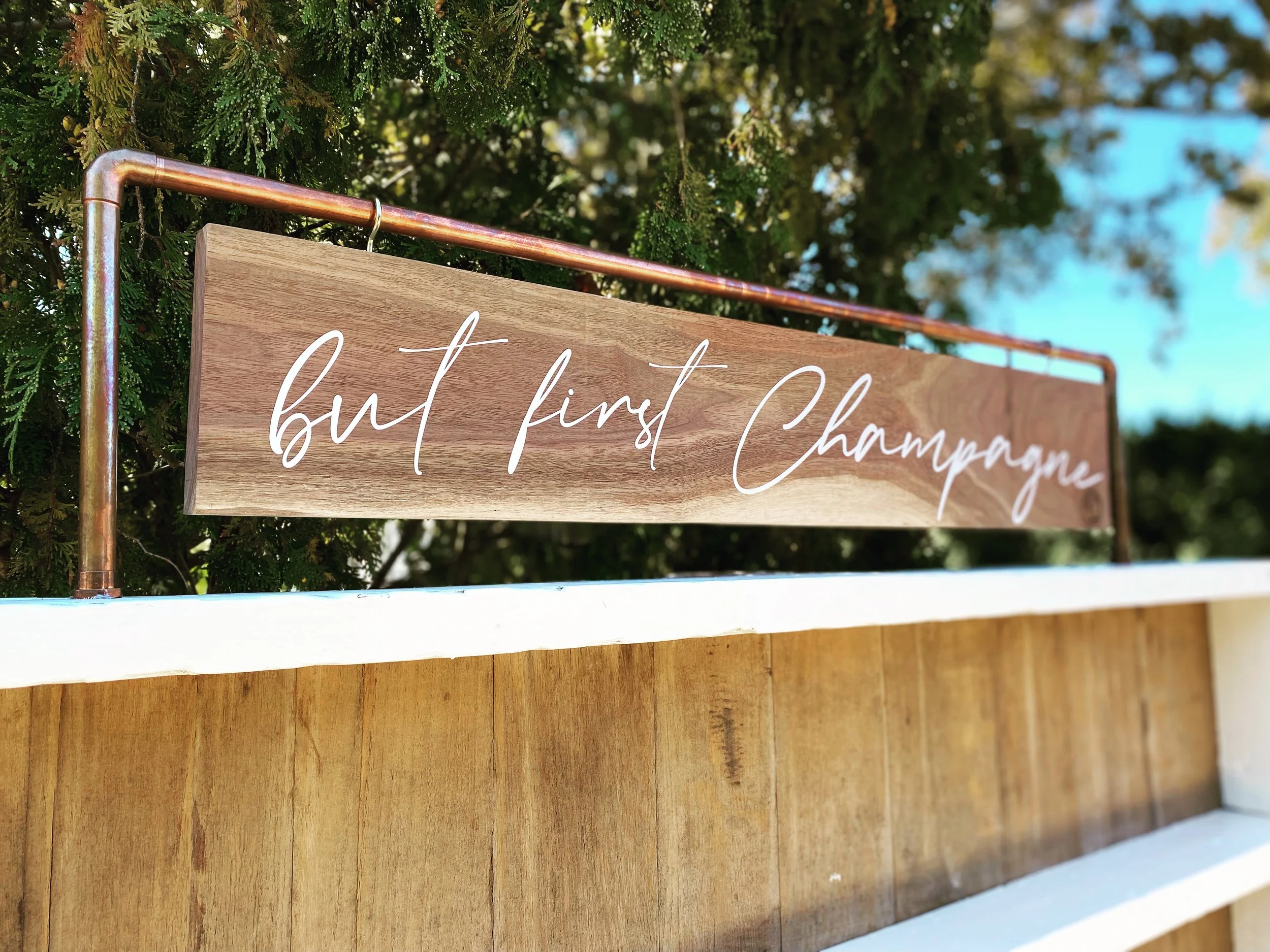 Wooden sign with "but first Champagne" in white script, mounted on a copper pipe frame, with green foliage in the background.
