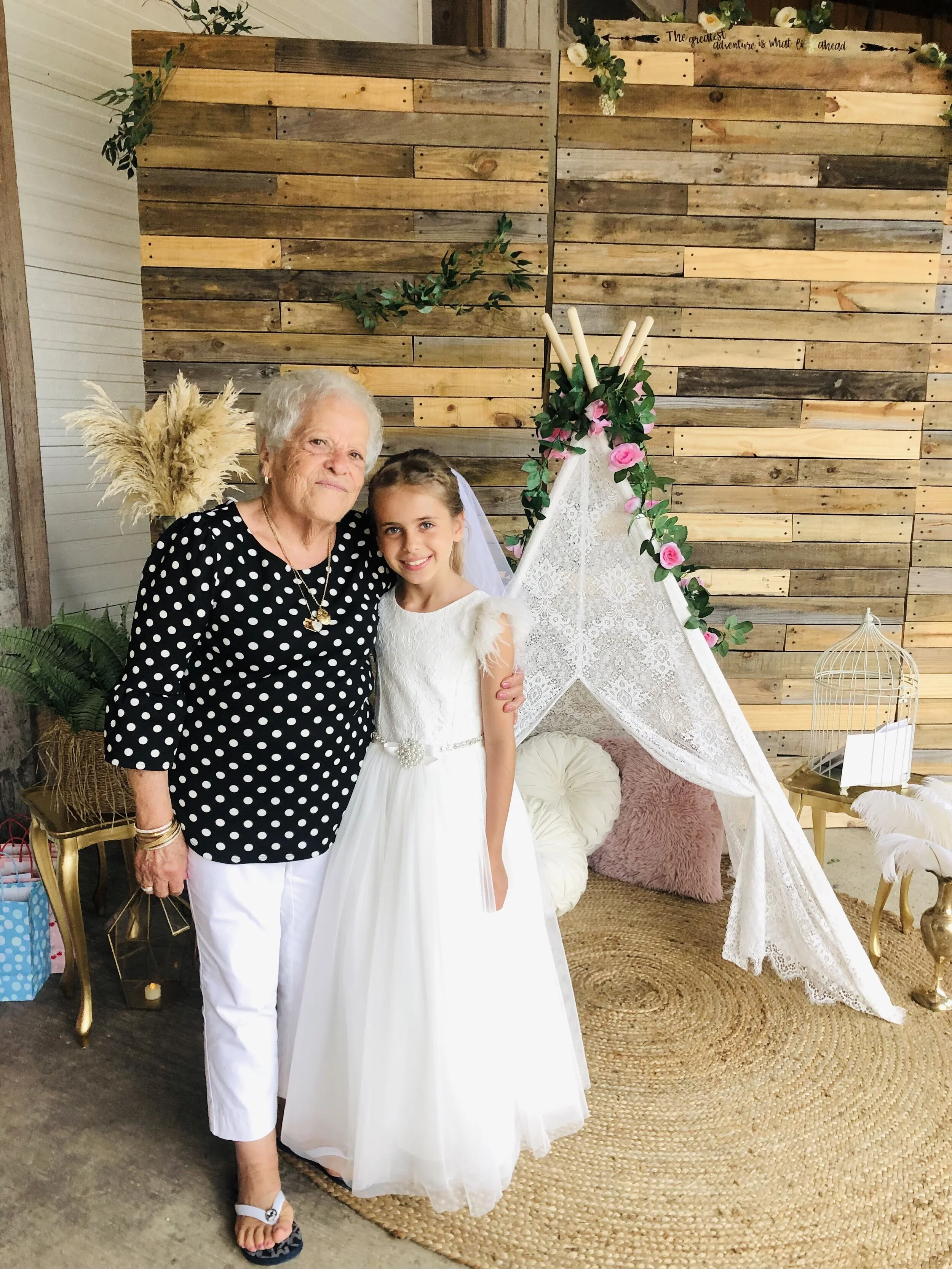 Older woman in black polka dot top and white pants standing next to a young girl in a white dress, posing in front of a rustic wooden backdrop with floral decorations and a lace tent.