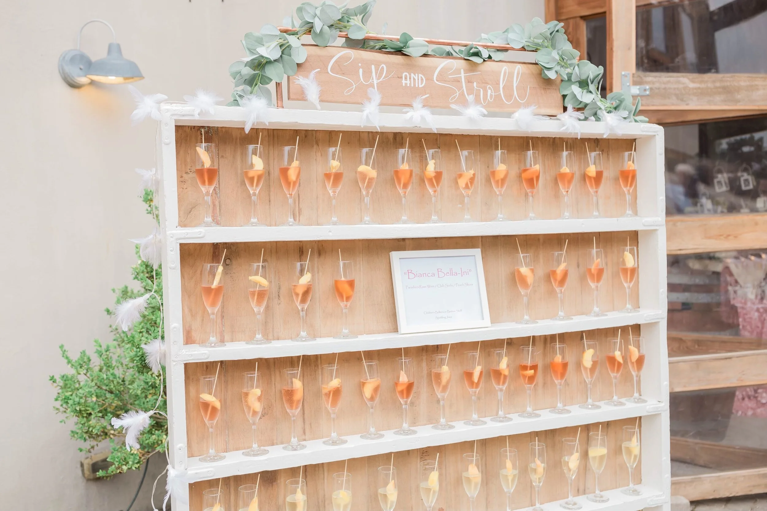 Wooden display shelf with rows of champagne glasses filled with pink drinks, garnished with fruit slices, labeled "Sip and Stroll."