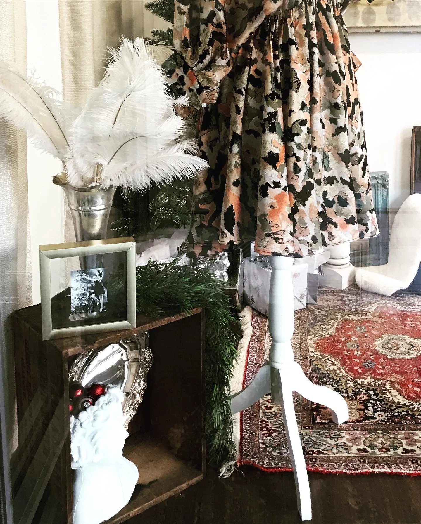 Mannequin with patterned dress, white feathers in a vase, framed photo, decorative garland, and an ornate mirror on a wooden box in a room with a red patterned rug.