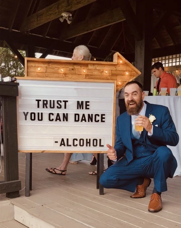 A man in a blue suit kneeling next to a lightbox sign that reads "Trust me, you can dance - Alcohol." He is holding a drink in one hand and smiling. The setting appears to be a casual indoor or outdoor gathering.