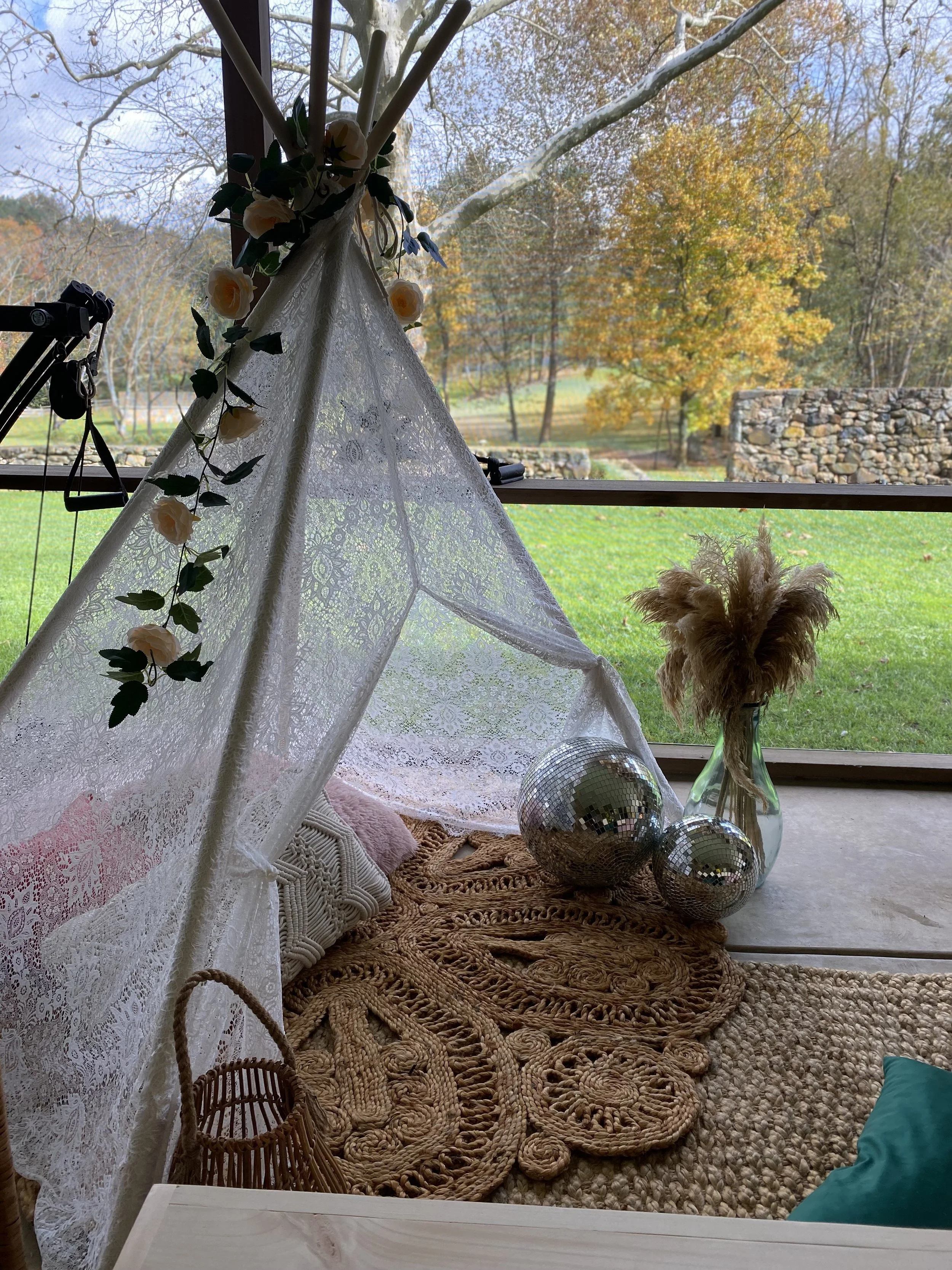 Decorative indoor teepee with lace fabric, adorned with flowers, set on a woven rug, featuring pillows, disco balls, and a vase of dried grass, positioned near a large window overlooking a garden.