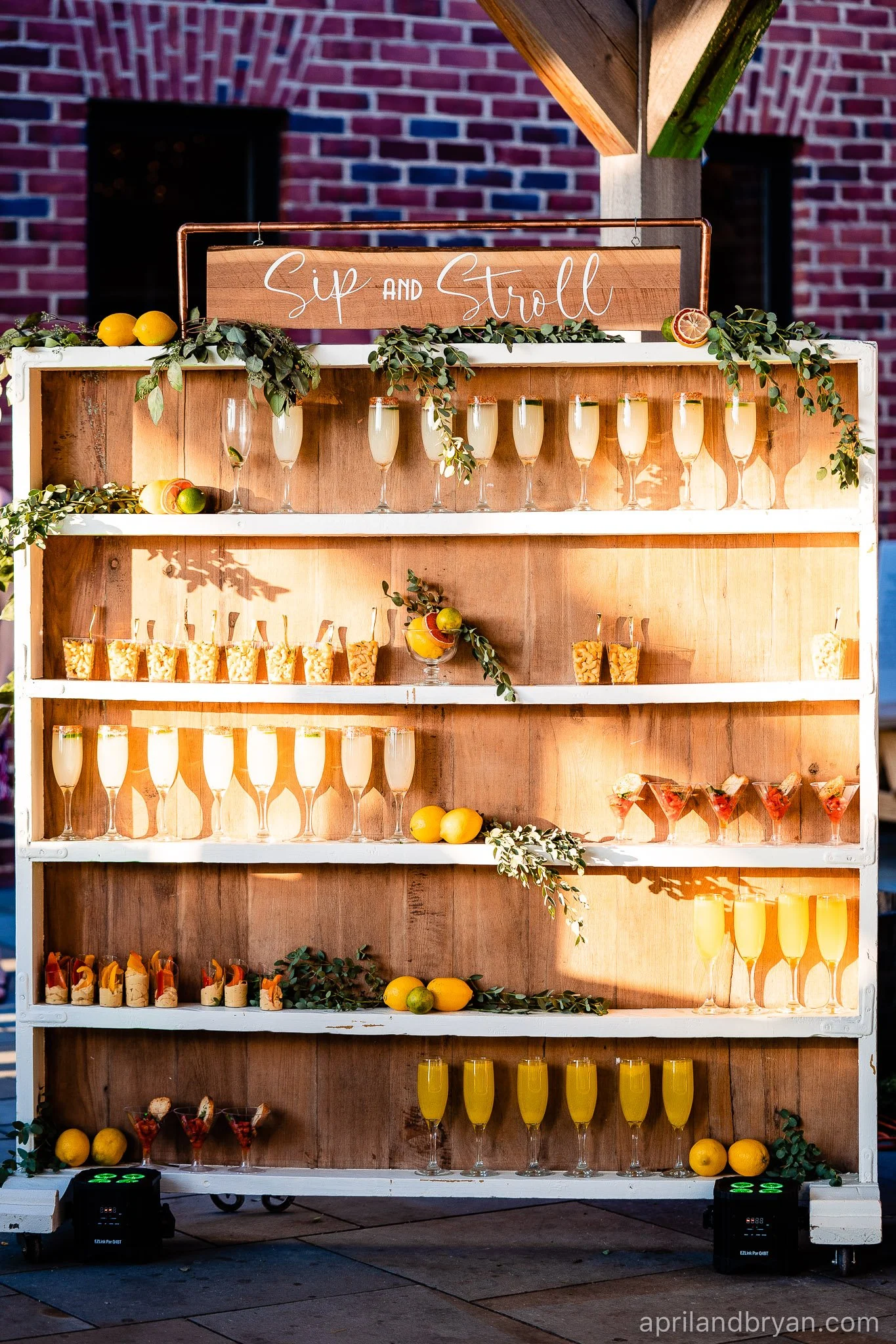 A wooden display shelf with multiple levels holding glasses of yellow and orange drinks, labeled 'Sip and Stroll.' The shelves are decorated with green foliage, citrus fruits, and small food items. The background features a brick wall.