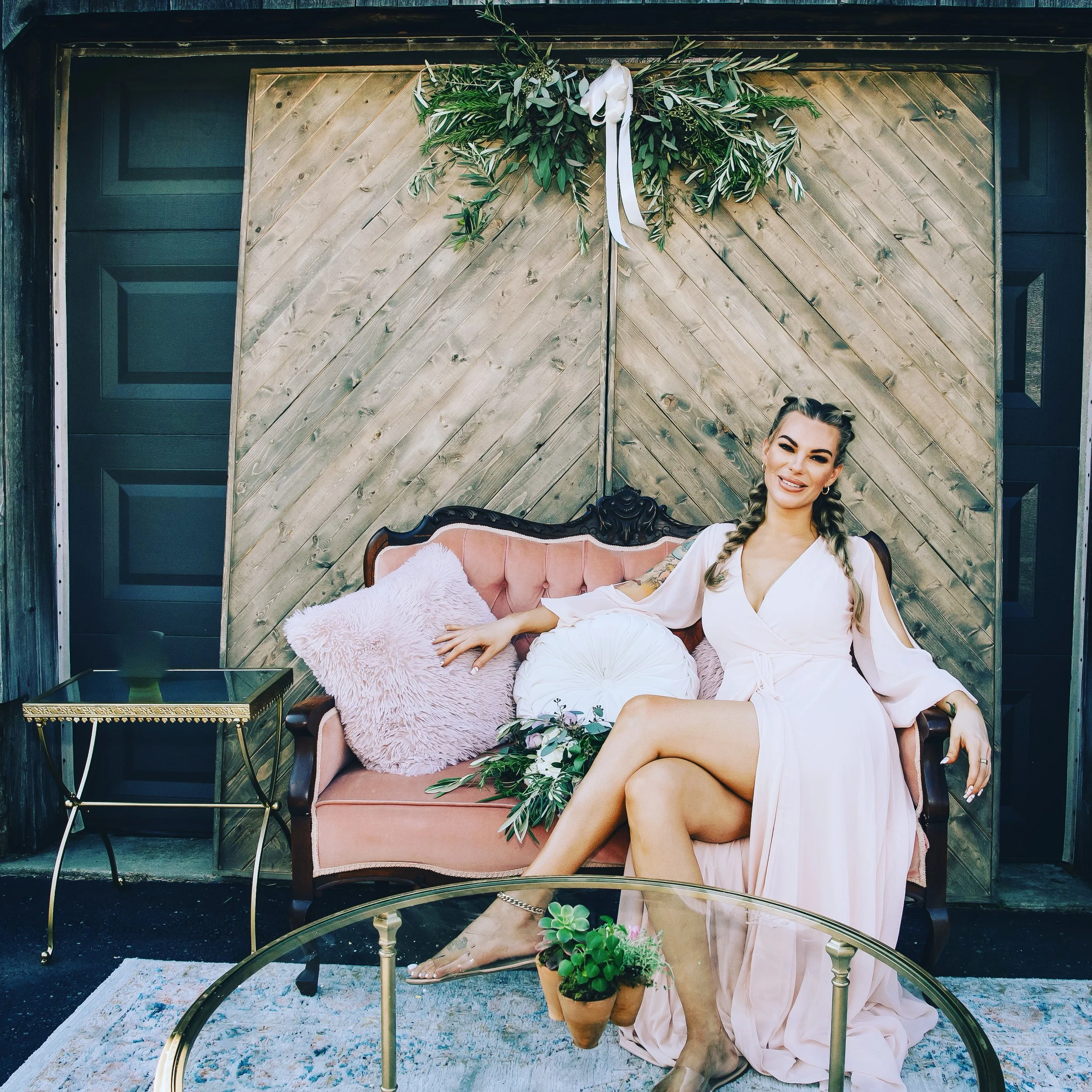 Woman in a light pink dress sitting on a vintage pink sofa with decorative pillows, surrounded by greenery and flowers, next to a glass coffee table with a small plant. The background features rustic wooden paneling with a green wreath and ribbon.