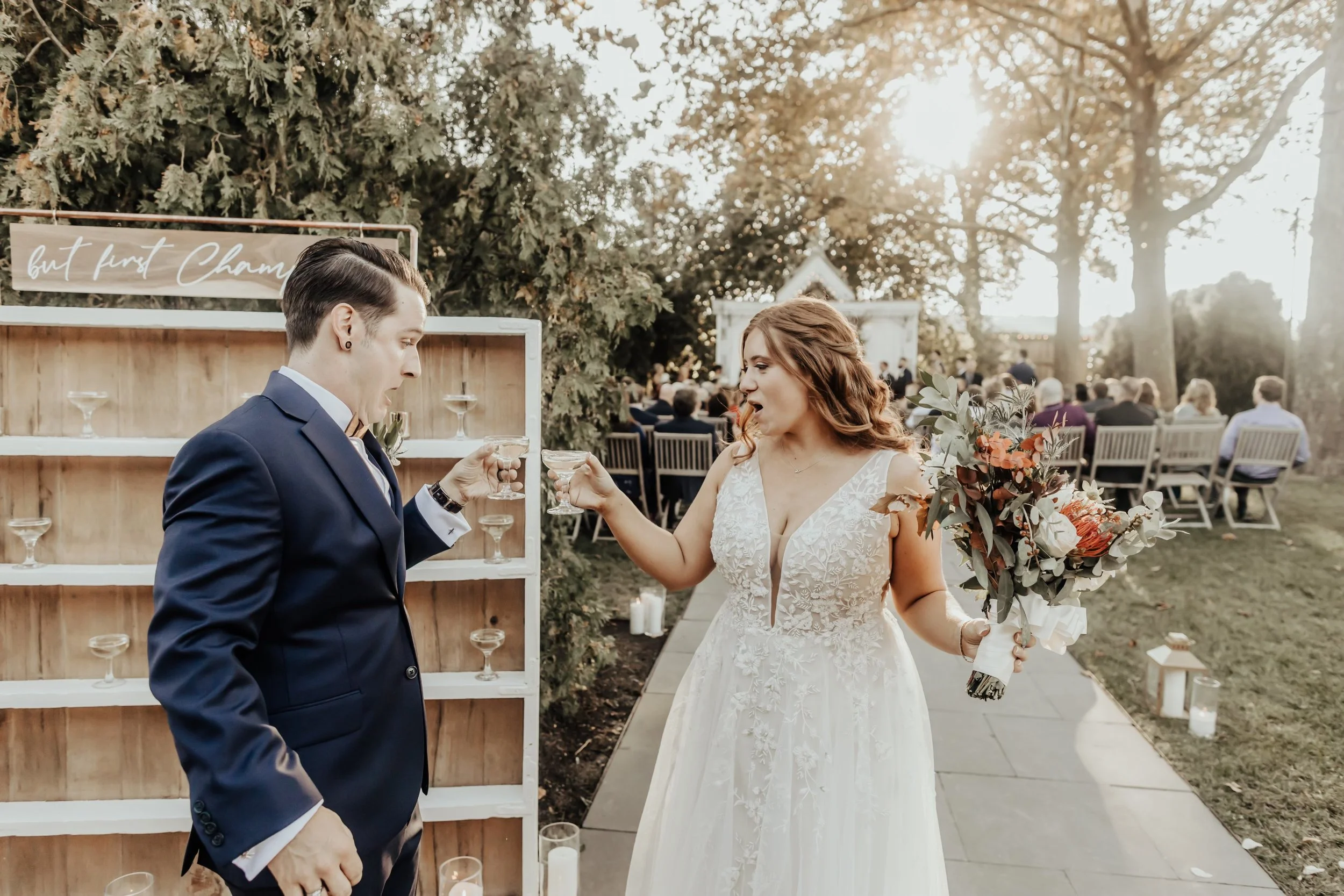 A bride and groom enjoying a champagne toast outdoors, with the bride holding a bouquet and wearing a lace wedding dress, and the groom in a blue suit. They're in front of a champagne display with a sign that says 'but first, champagne,' surrounded b