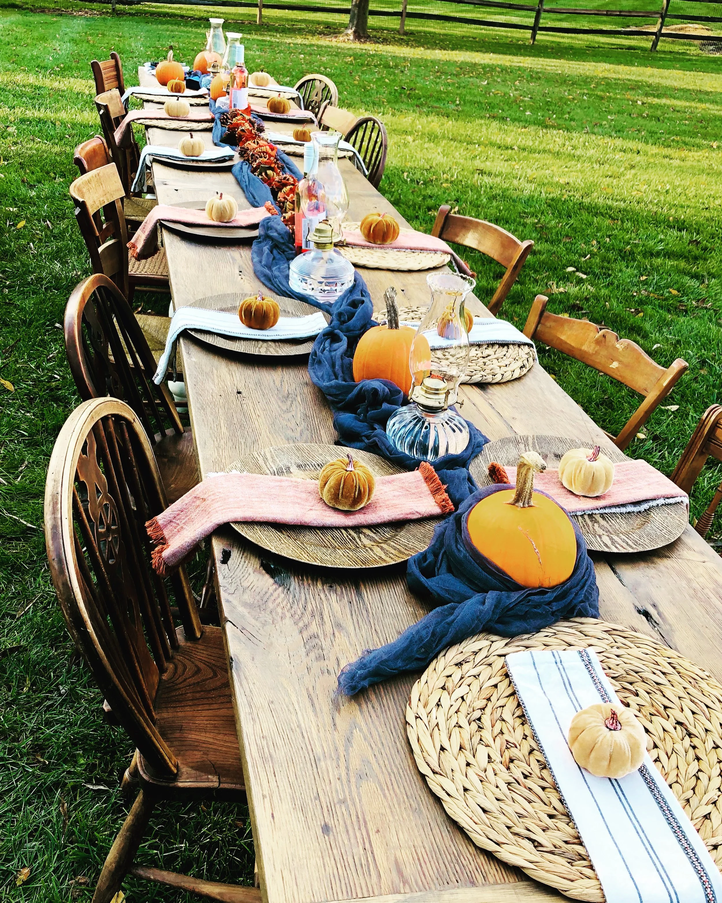 Outdoor dining table decorated for fall with pumpkins, table settings, and centerpieces on a grassy lawn.