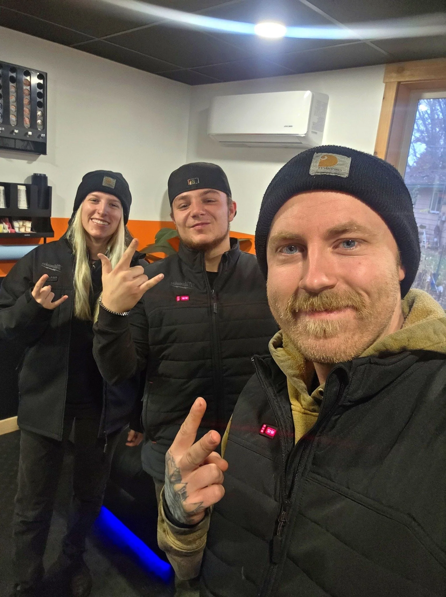 Three people taking a selfie indoors, all wearing black beanies and jackets, making a rock sign with their hands. The background shows a window, an air conditioning unit, and shelves with supplies.