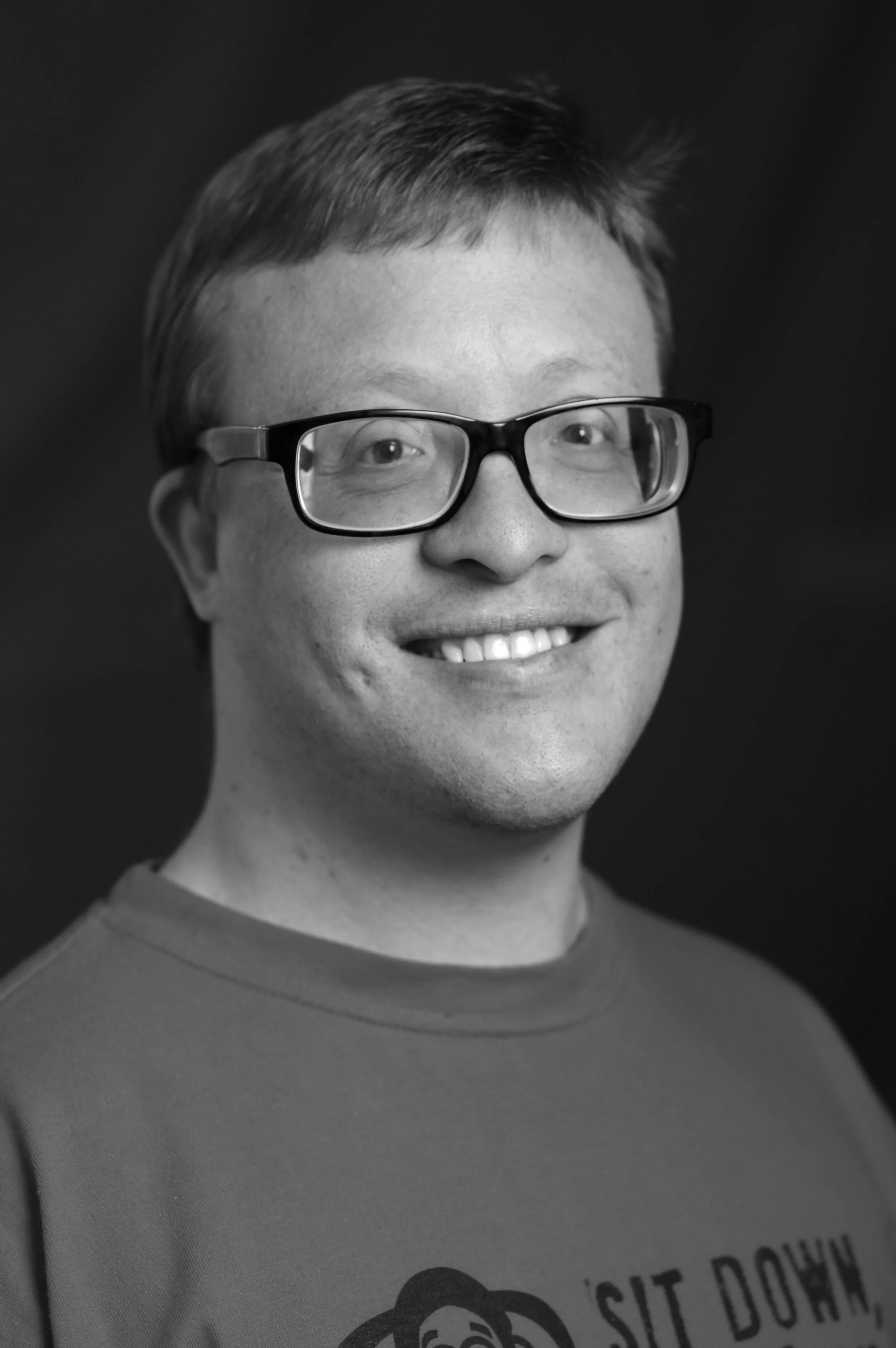 Black and white photo of James, he is wearing black rimmed glasses, has short blond hair and is smiling at the camera.