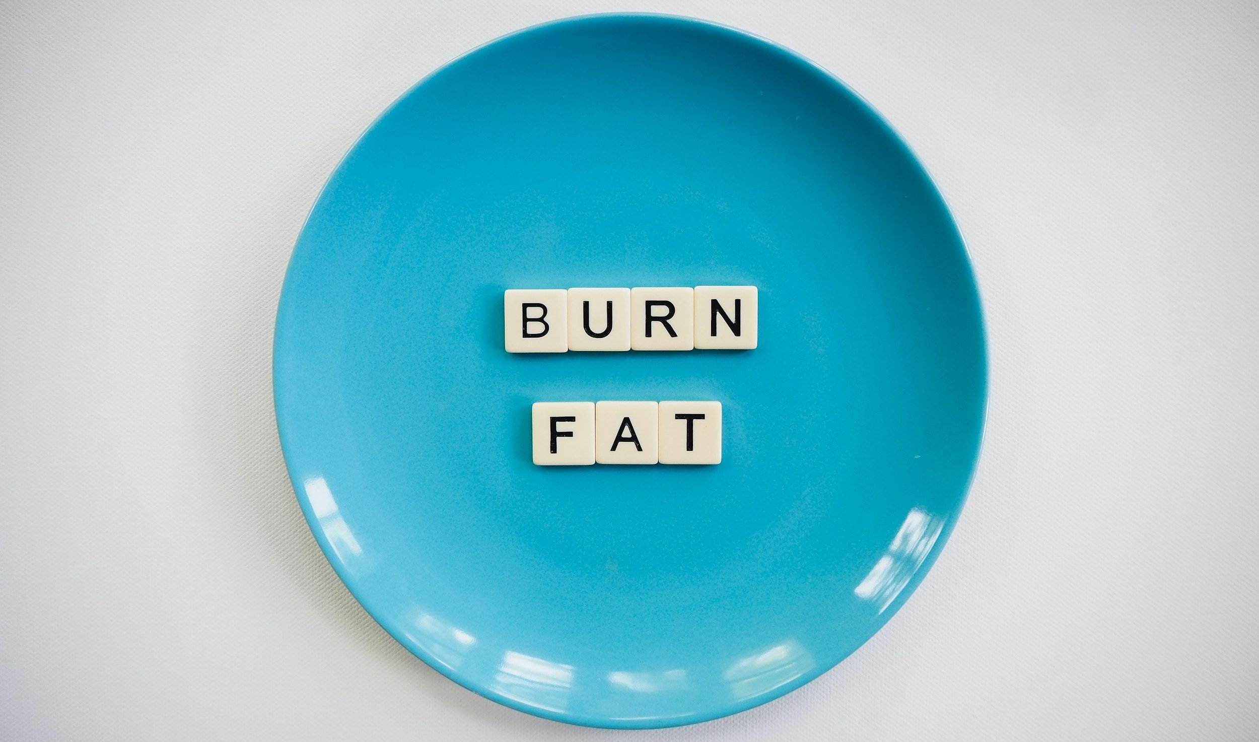 Blue plate with scrabble tiles spelling 'BURN' and 'FAT' on a white textured background.