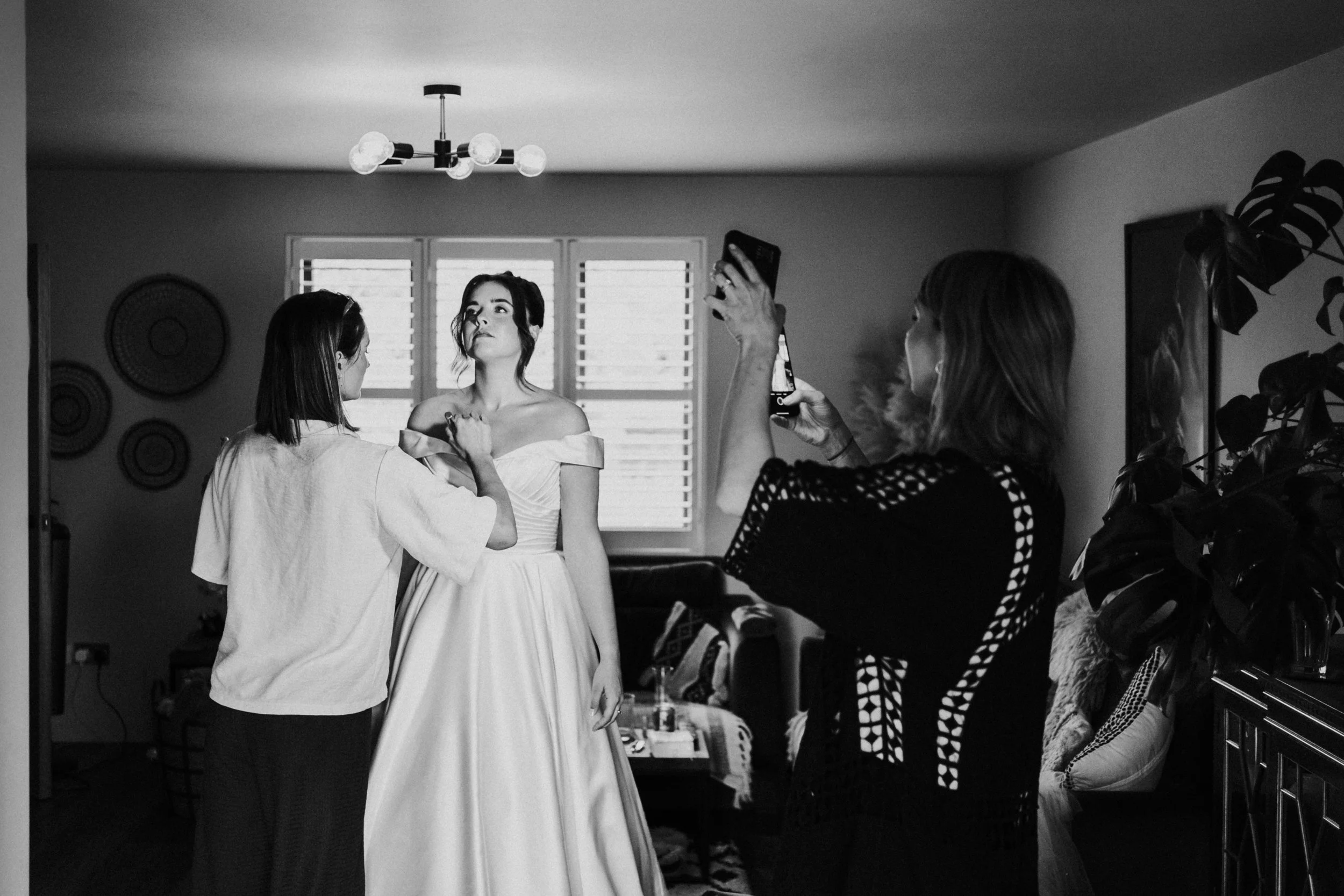 Bride standing having her makeup touched up by artist with assistant holding mirror before a Worton Kitchen Garden, Oxfordshire Wedding